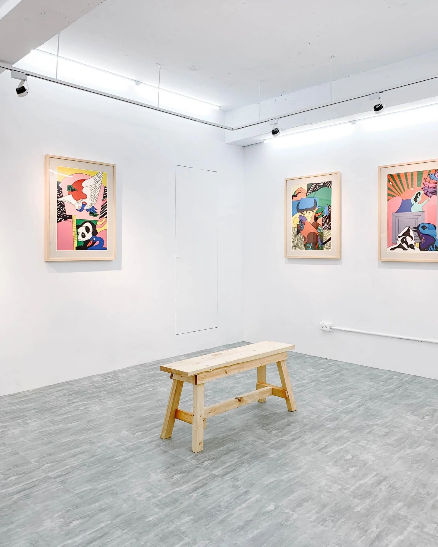 A minimalistic art gallery with three colorful, abstract framed artworks on white walls, a wooden bench in the center, and a technical ceiling with track lighting.