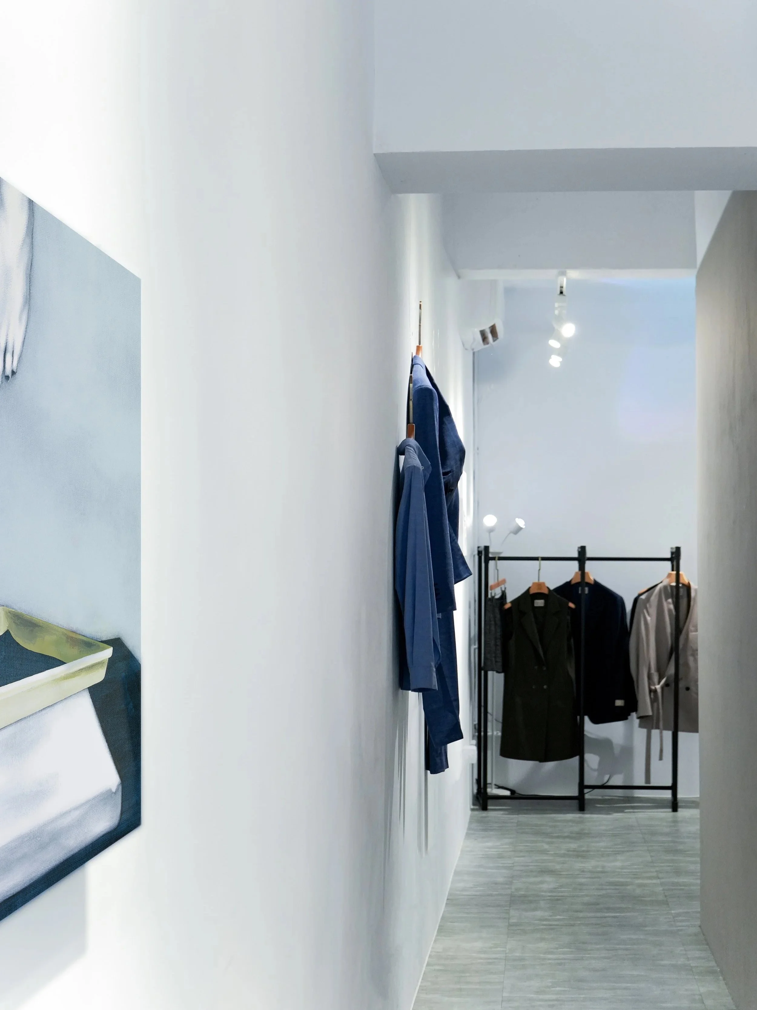 A minimalist retail clothing store with a white wall, a black clothing rack displaying suits, and a clothing rack with jackets and shirts in a bright, well-lit space.