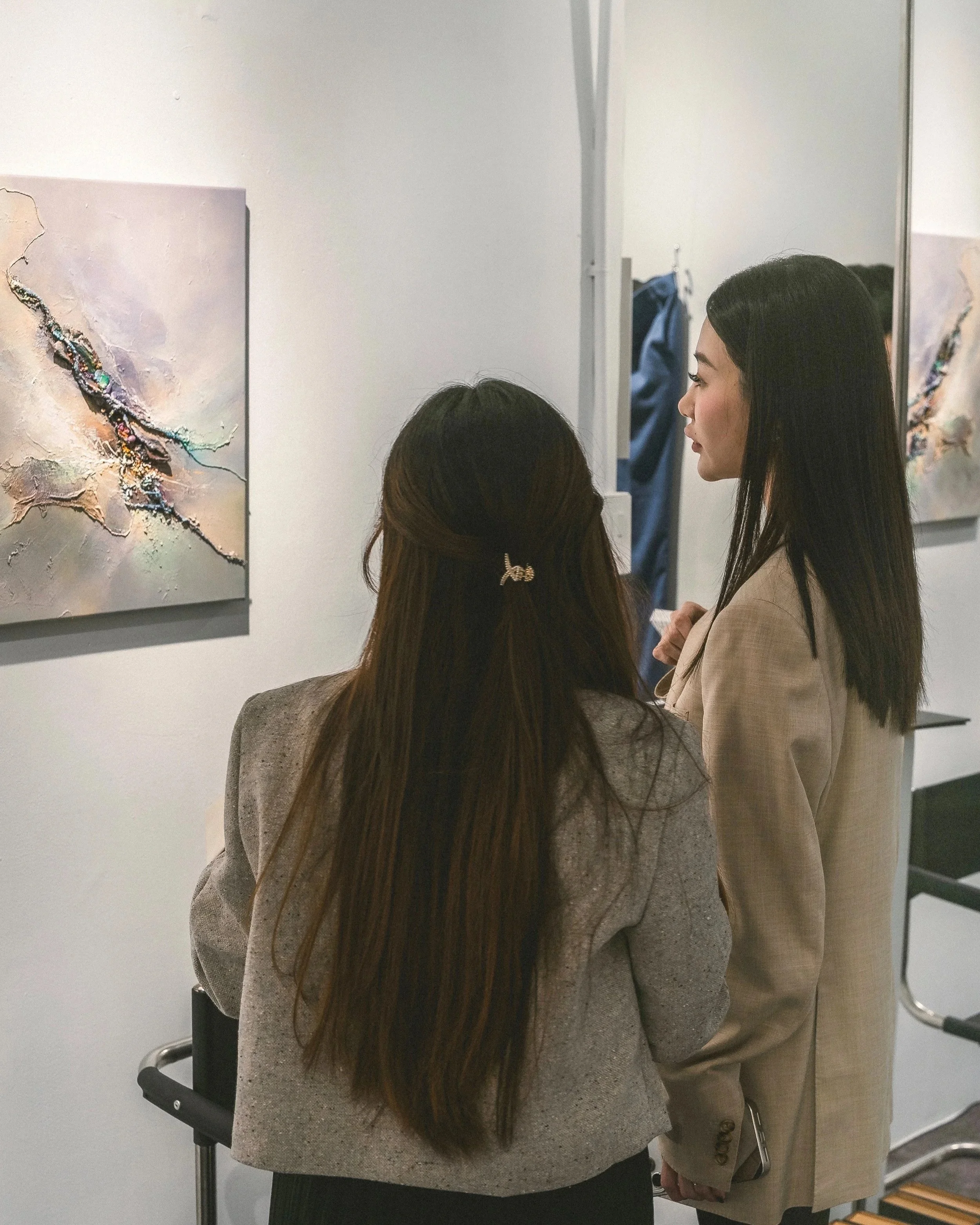 Two women stand in an art gallery looking at a painting on the wall. One woman has long brown hair with a hair clip, dressed in a beige or gray blazer. The other woman has long black hair and is wearing a light-colored blazer. There is a mirror refle