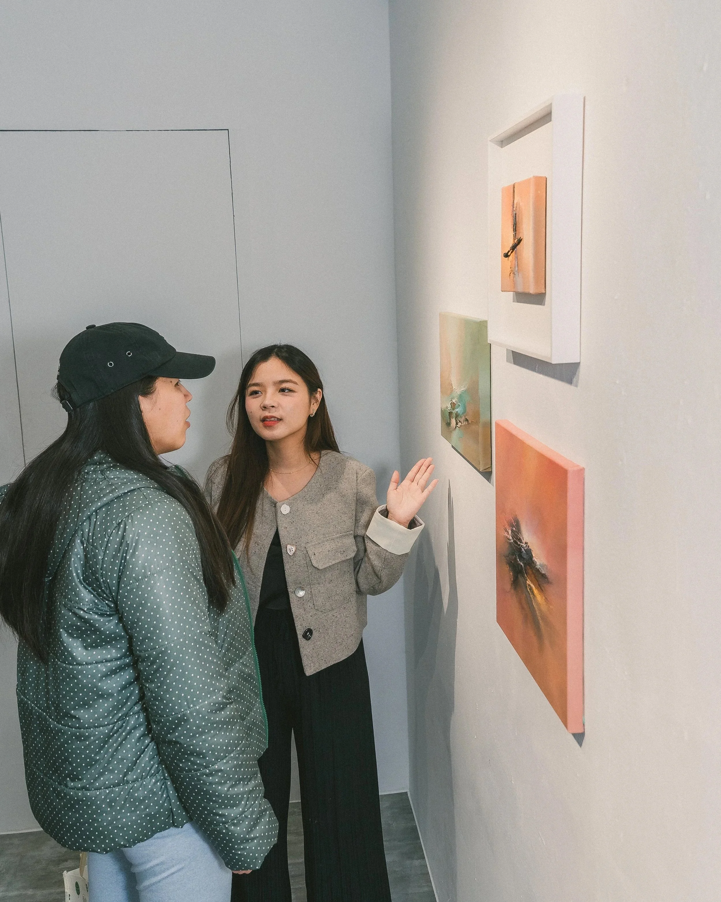 Two women talking in an art gallery, with various paintings on the wall.