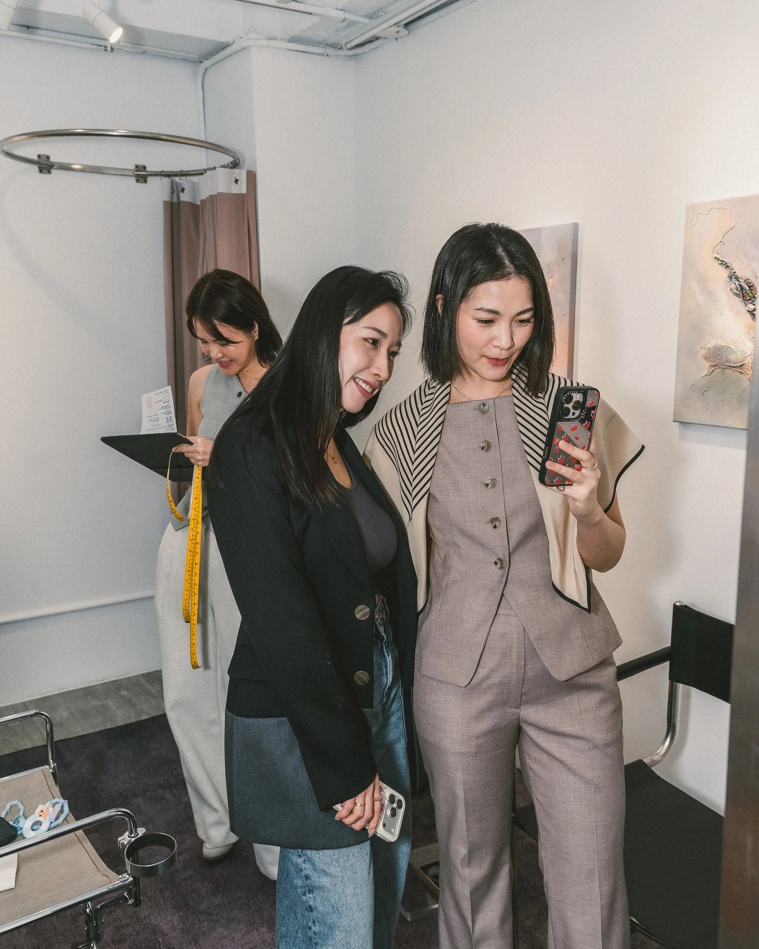 Three women standing together at an art gallery, looking at a phone. One woman is in a light-colored blazer with striped collar, another in a black blazer, and the third in a beige blazer. Artwork is displayed on the wall behind them, and a seating a