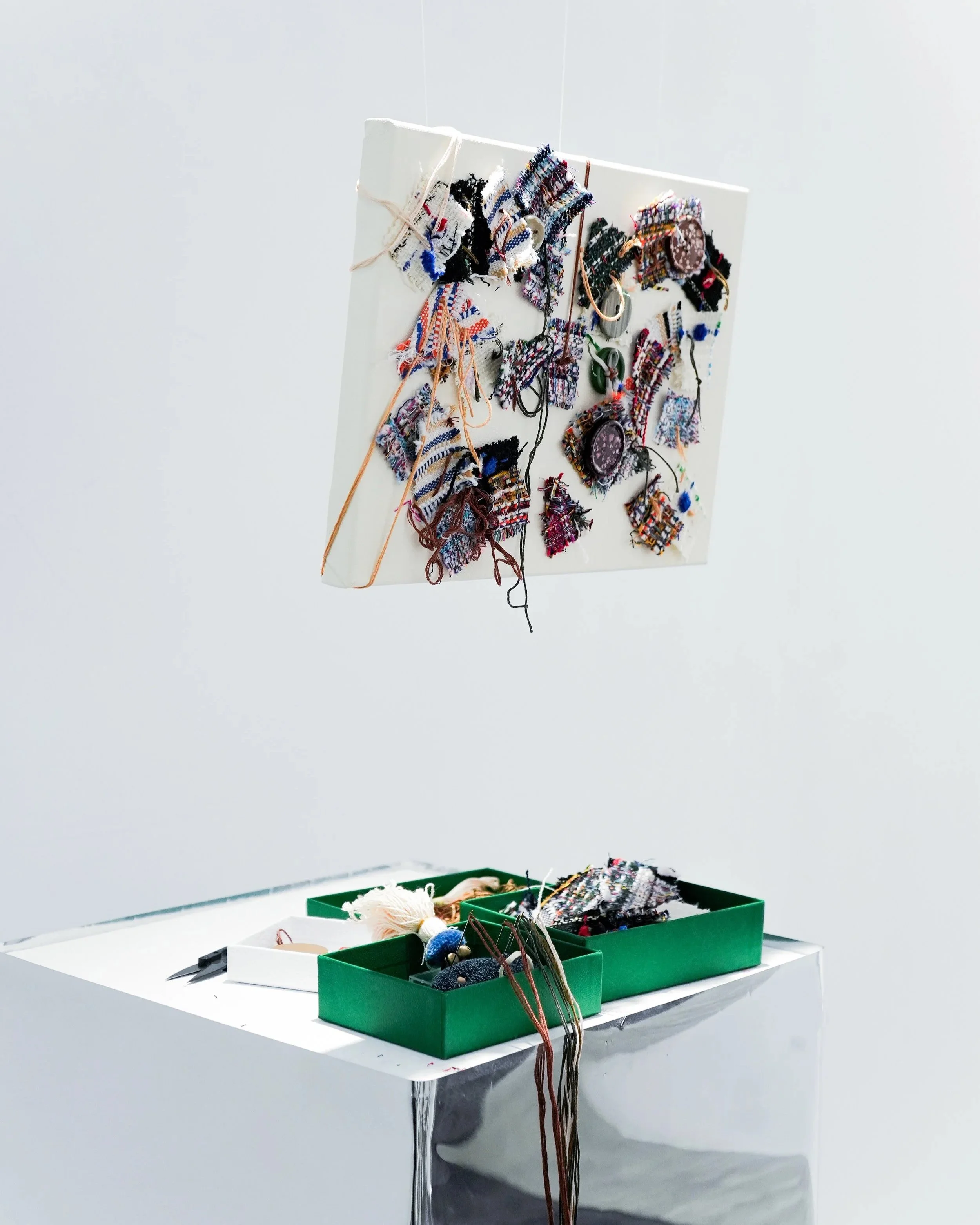 An art installation features a white canvas hanging on a wall, decorated with various colorful woven textile fragments attached with strings. A green box filled with yarn and more textile pieces is placed on a white surface below the canvas.