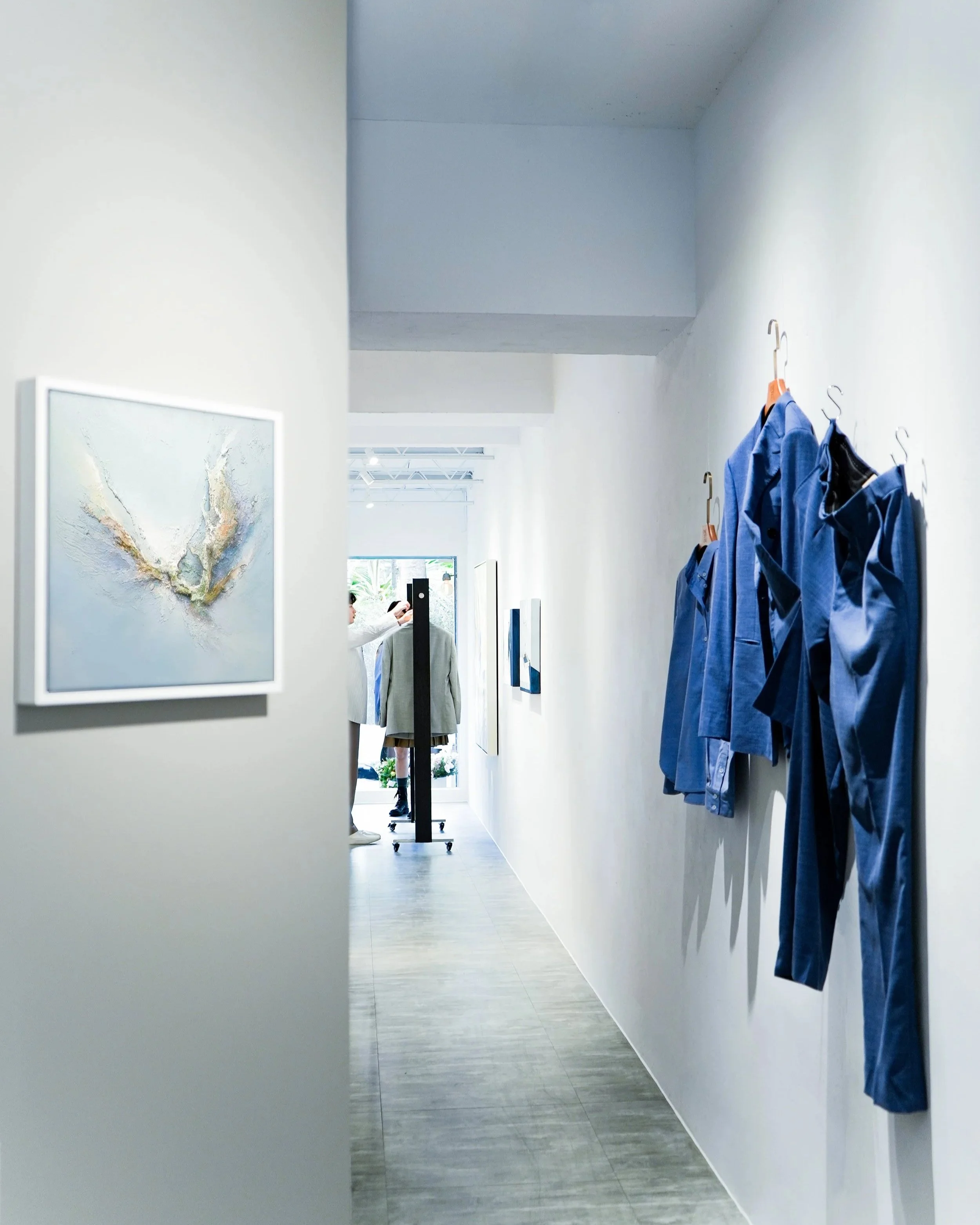 Hallway with blue shirts hanging on hooks on the right wall, artwork on the left wall, and a person in the background using a magnifying glass.