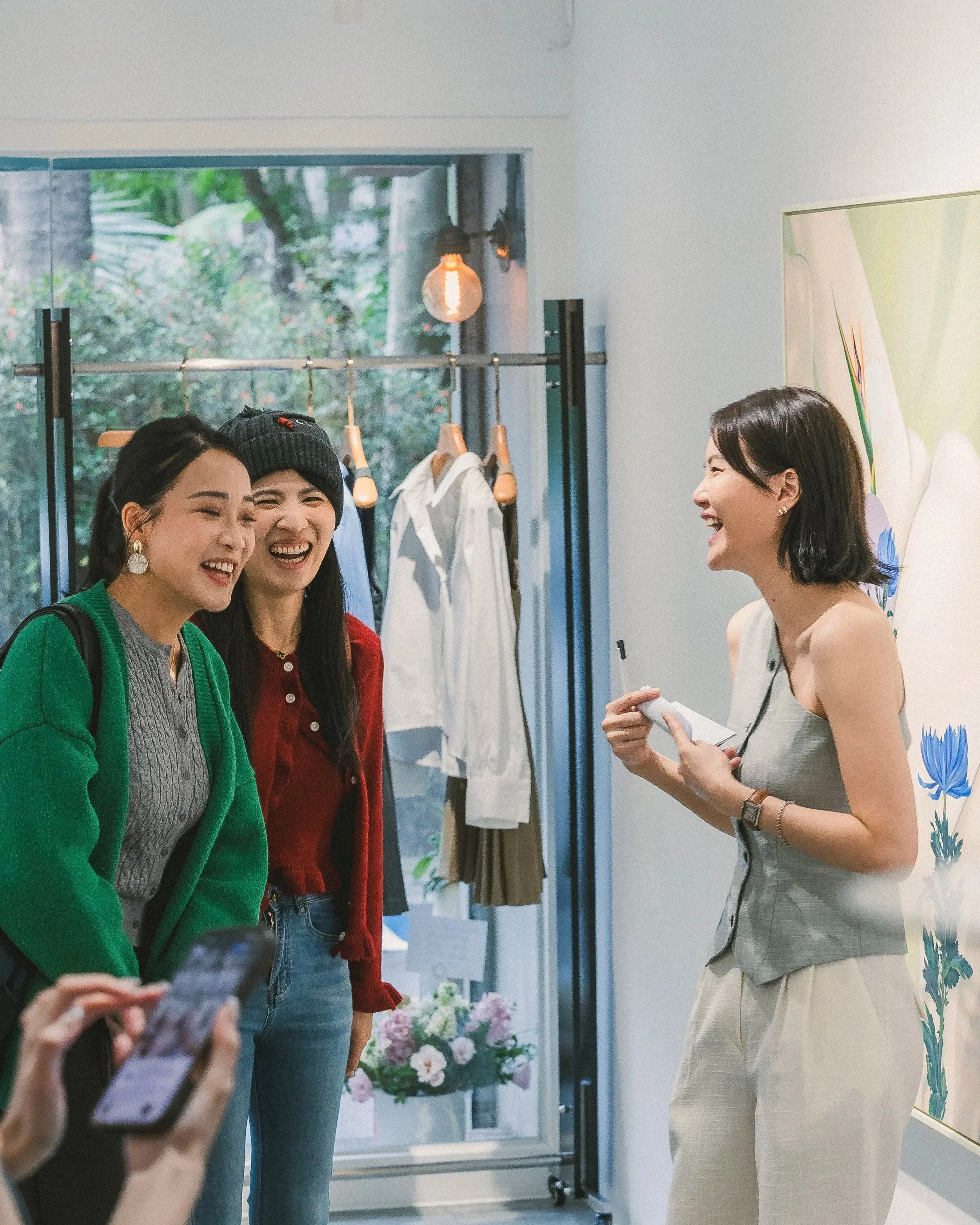 Three women are standing indoors, engaged in conversation and smiling. One woman on the right is holding a notepad and pen. Two women on the left are wearing casual clothes, one with a green cardigan and another with a red sweater. The background fea