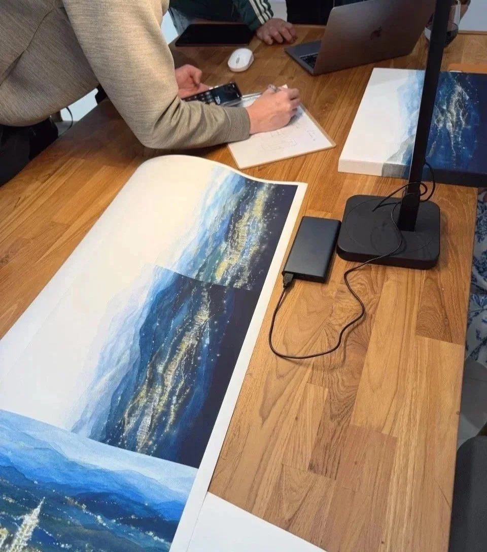 People working on a detailed landscape painting of mountains and a starry night, with art supplies, laptops, and a monitor on a wooden table.
