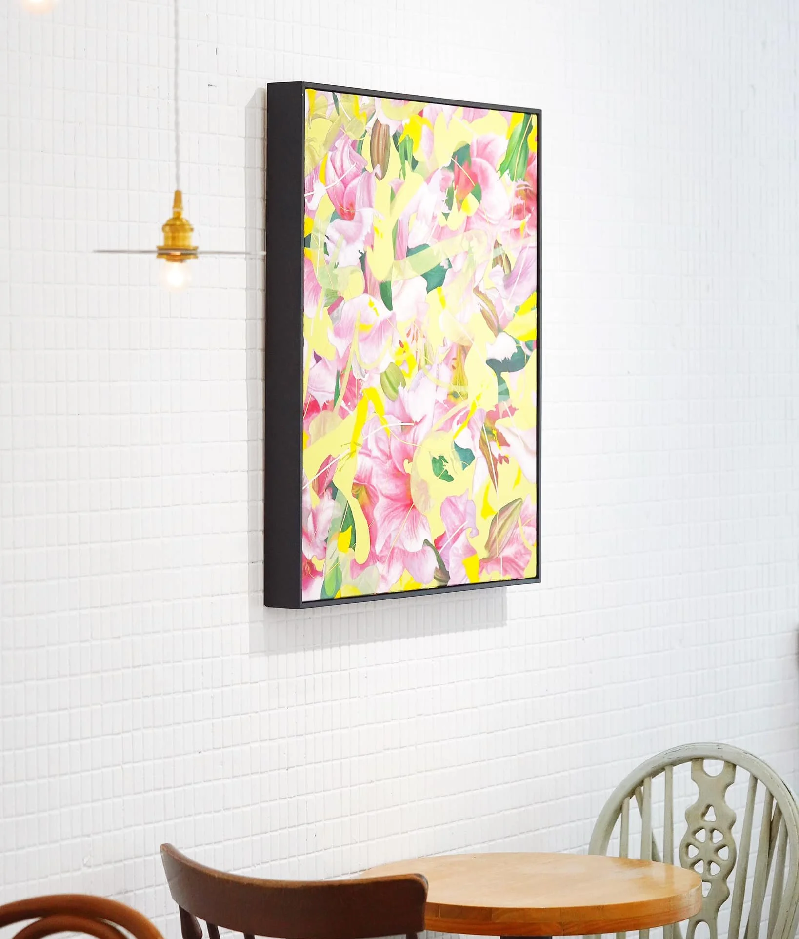 Colorful abstract art painting hanging on a white tiled wall, adjacent to a small hanging light bulb fixture. Part of a dining area with a wooden table and mismatched chairs is visible.