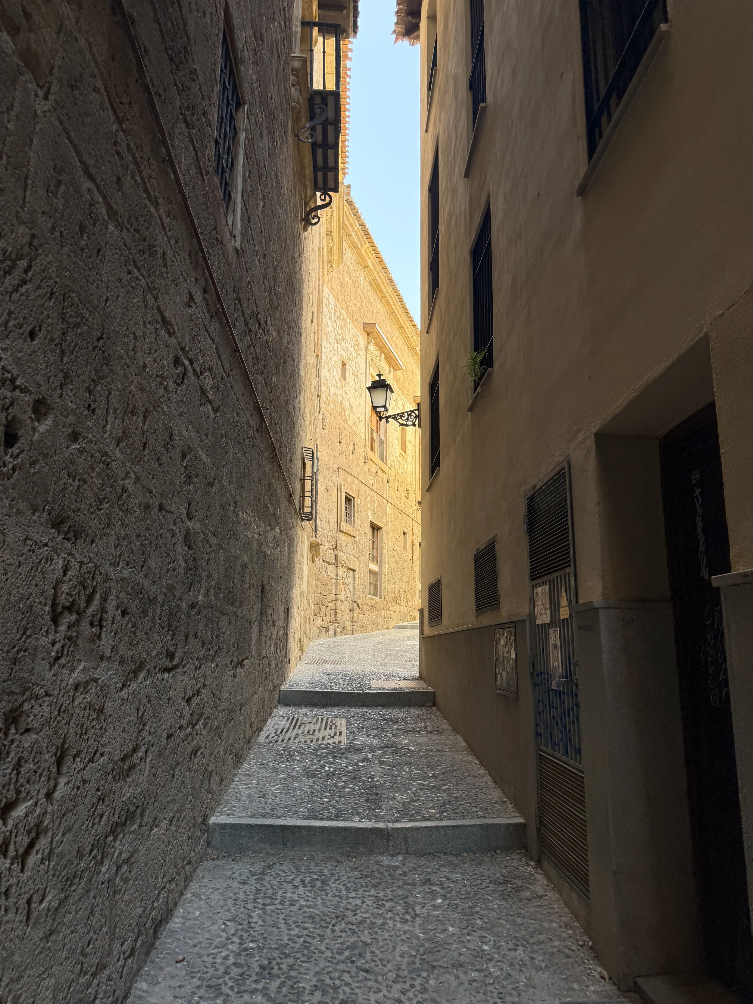 A narrow alleyway between stone and plaster buildings, with steps leading up and a lantern hanging on the wall.