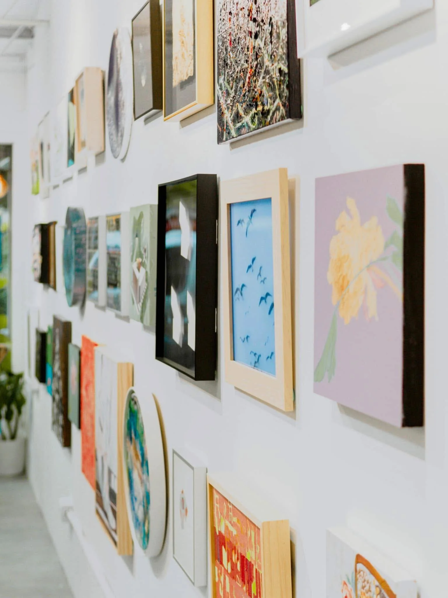 A white wall displaying multiple framed paintings and art pieces in various sizes and styles, arranged in a grid-like pattern.