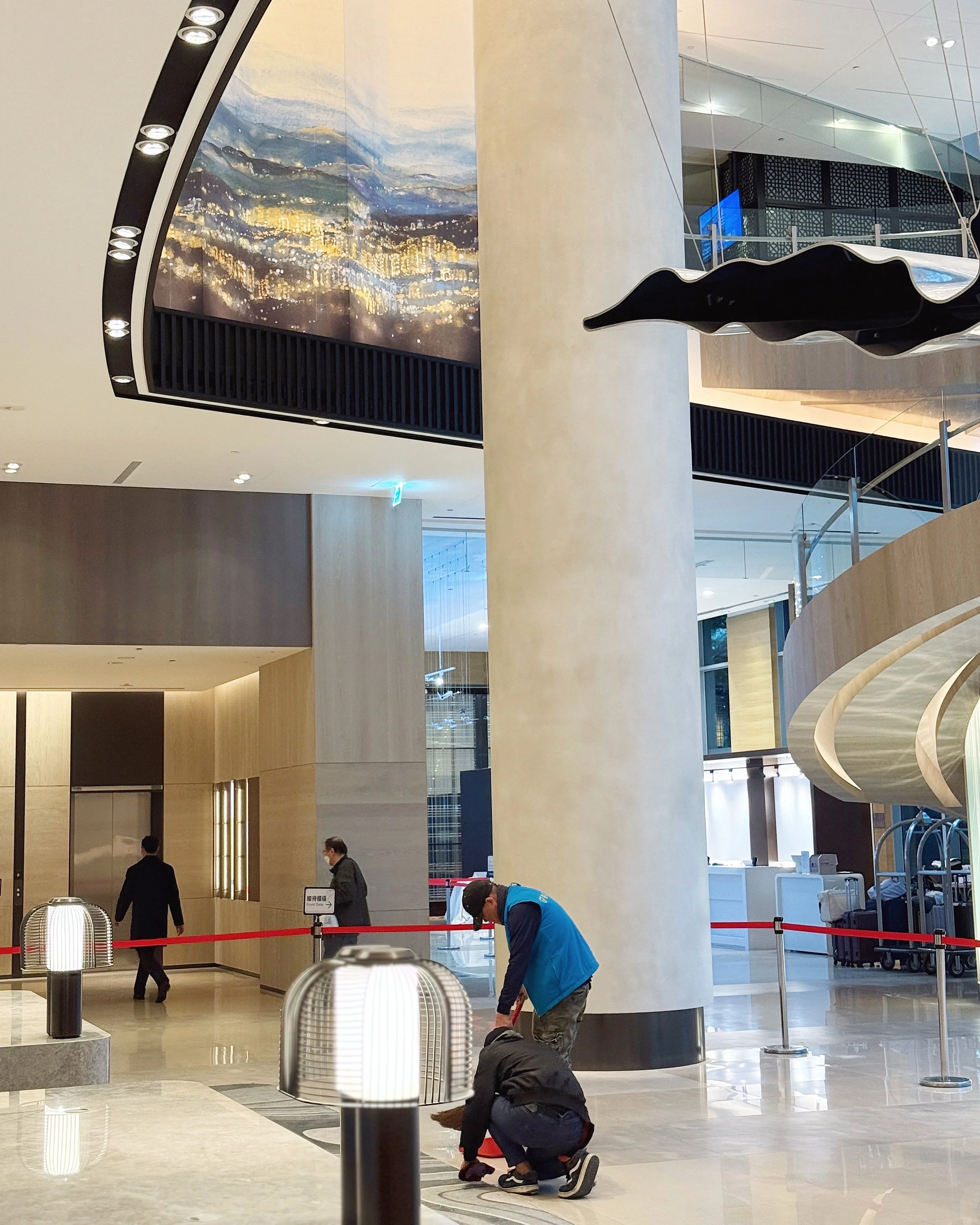 People working at an airport or train station, possibly cleaning or preparing the area. The space is spacious and modern with bright lighting, large pillars, and decorative elements like a large cityscape mural and architectural features.
