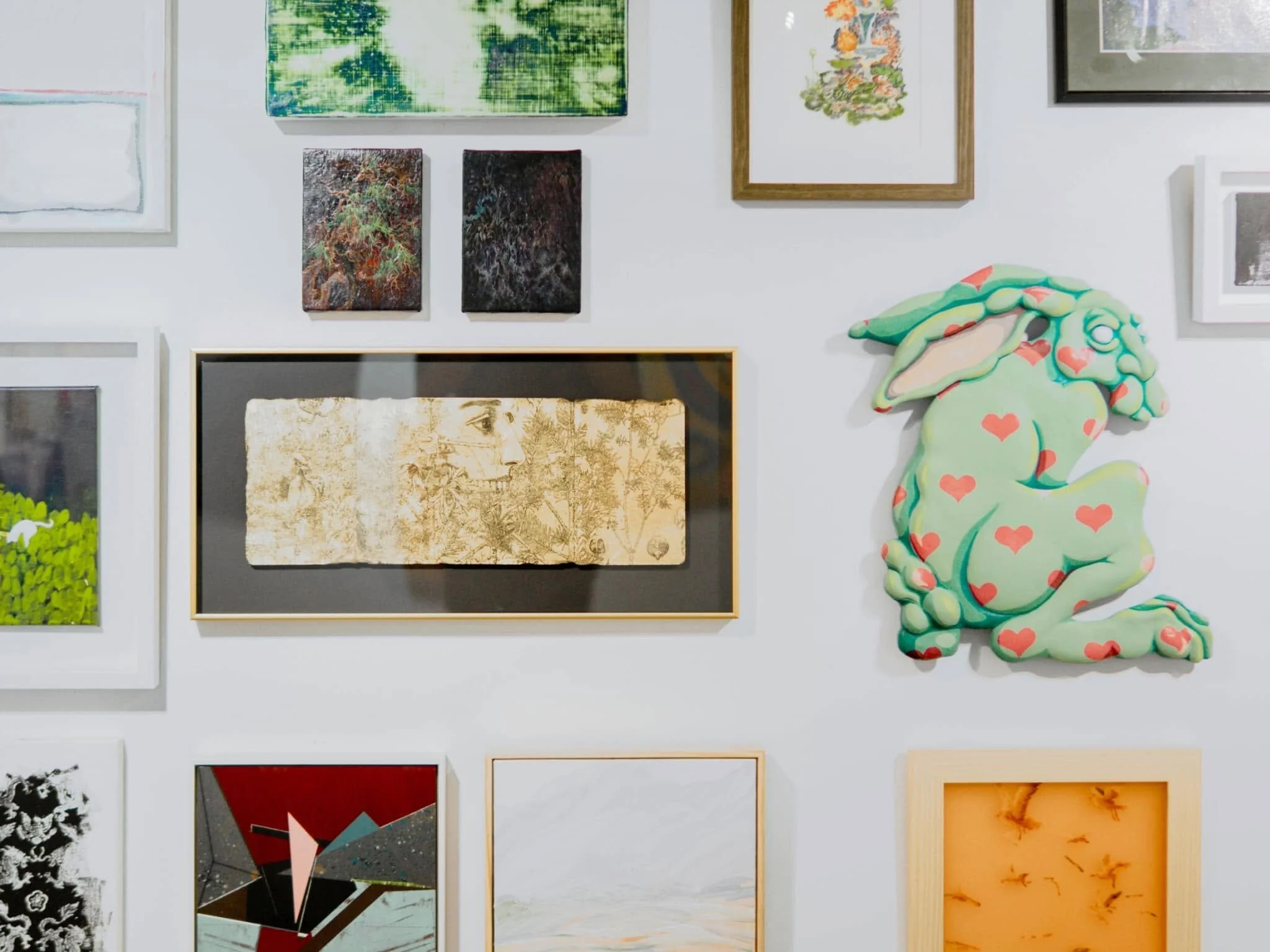 A gallery wall with various framed artworks, including abstract paintings, a portrait of a face, and a colorful stuffed bunny with heart patterns.