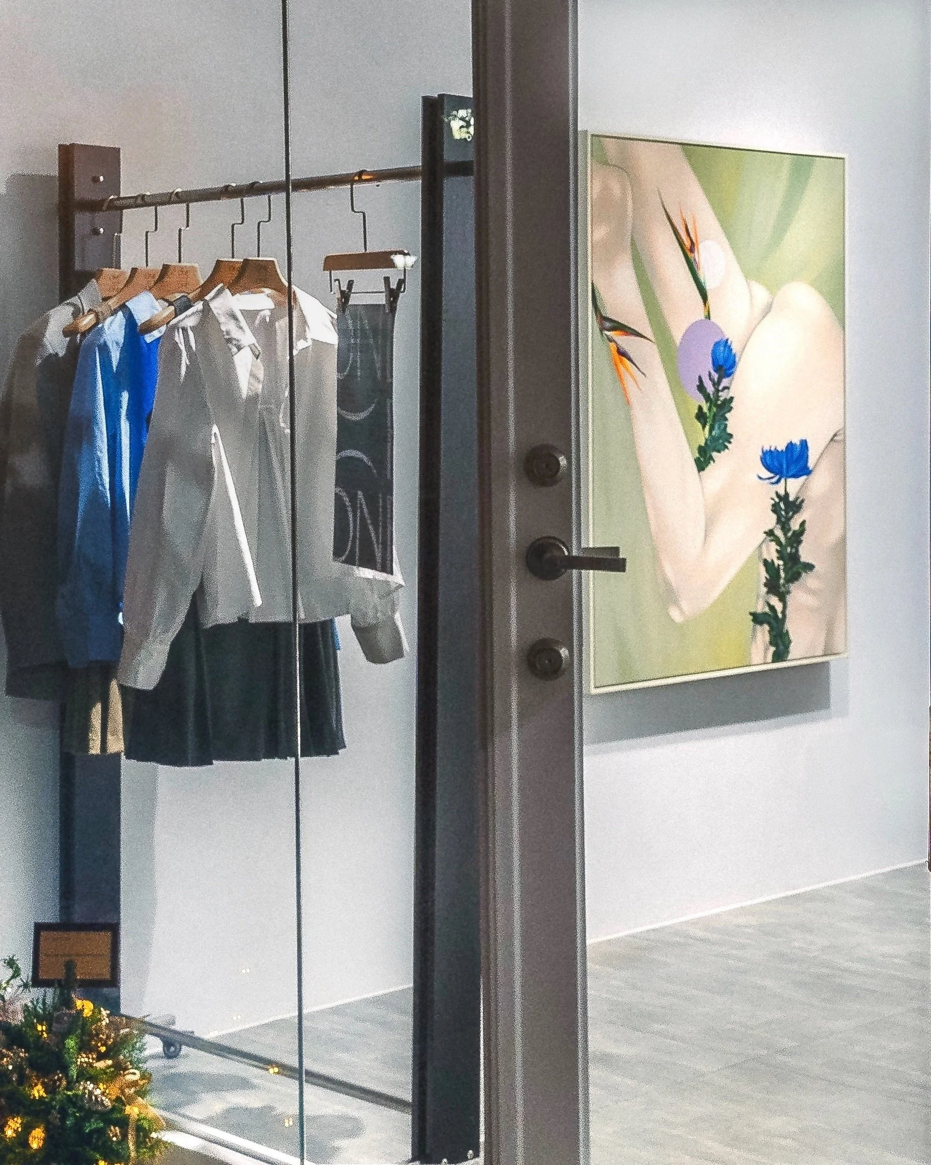 Clothes hanging on a rack inside a store, viewed through a glass door with a partial reflection of a painting of a hand with flowers.
