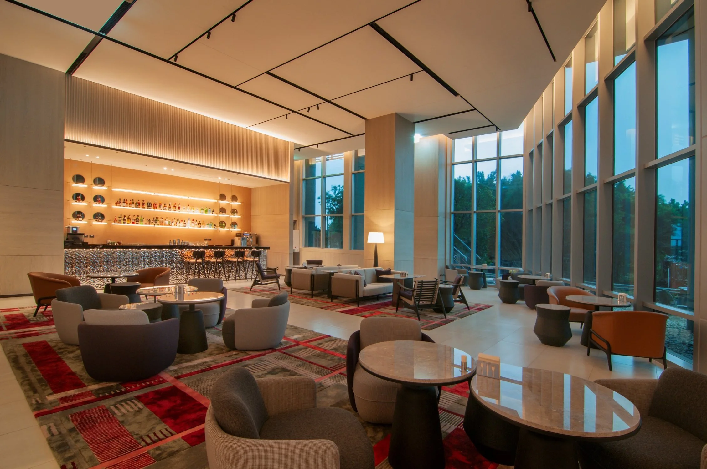 Interior of a modern lounge or bar area with large floor-to-ceiling windows, contemporary furniture, and a bar with bottles on display. The ceiling features geometric black lines and warm lighting, and there is a patterned red and gray rug on the flo