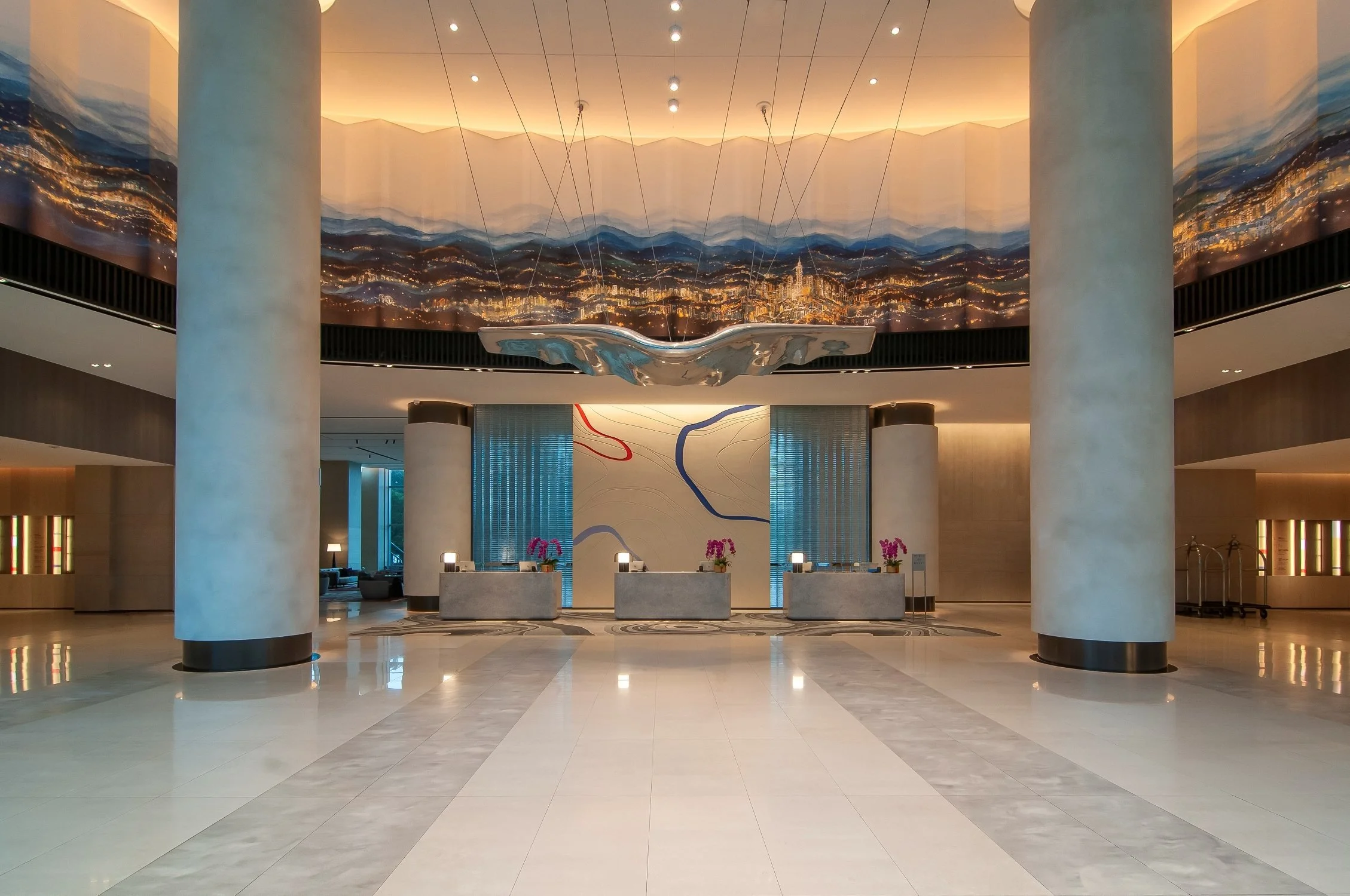 Hotel lobby with polished marble floors, large white columns, a reception desk with flower arrangements, and a large artistic cityscape mural on the ceiling.