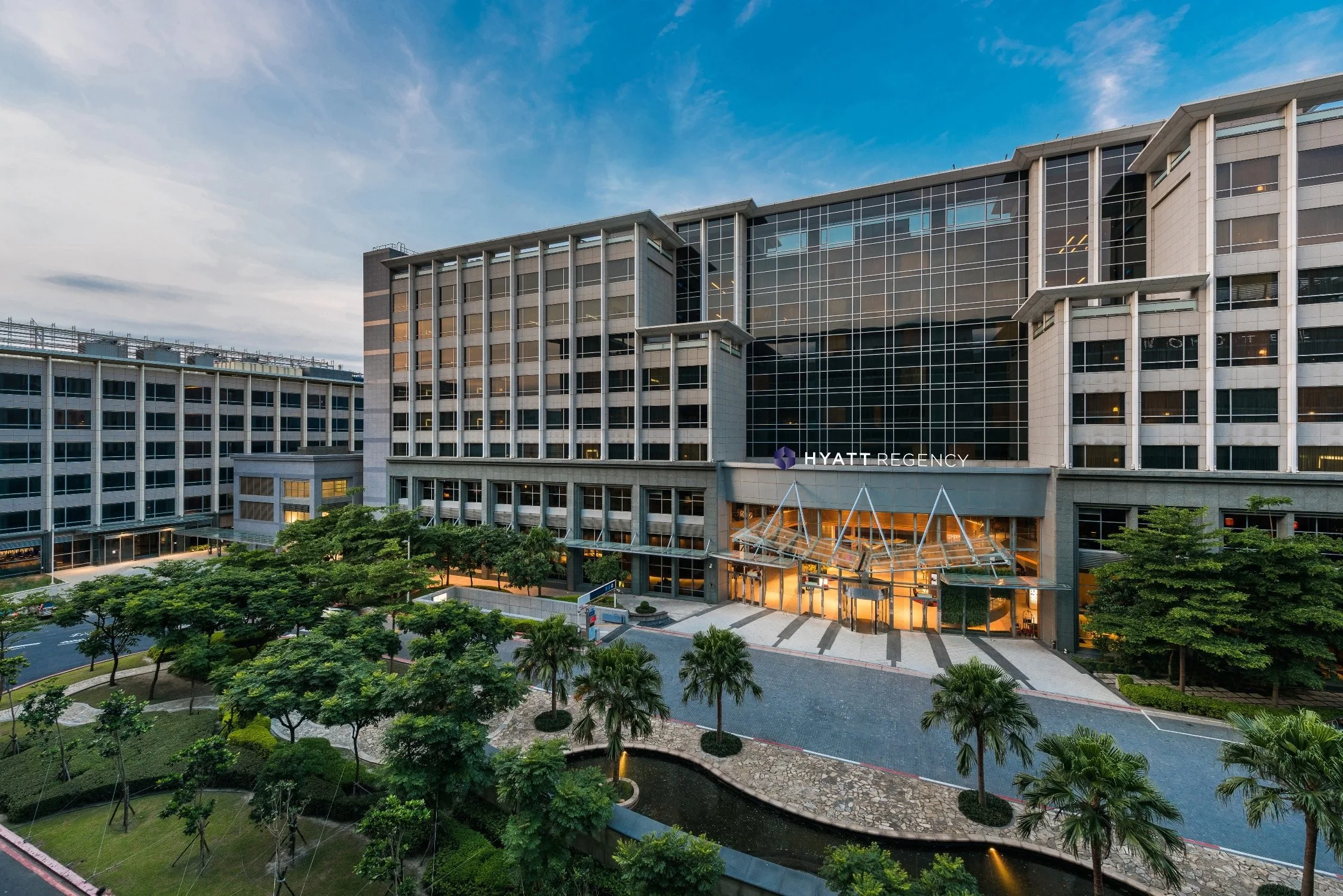 Exterior view of the Hyatt Regency hotel at dusk, featuring a modern multi-story building with large glass windows, surrounded by trees and a landscaped area with a pond.