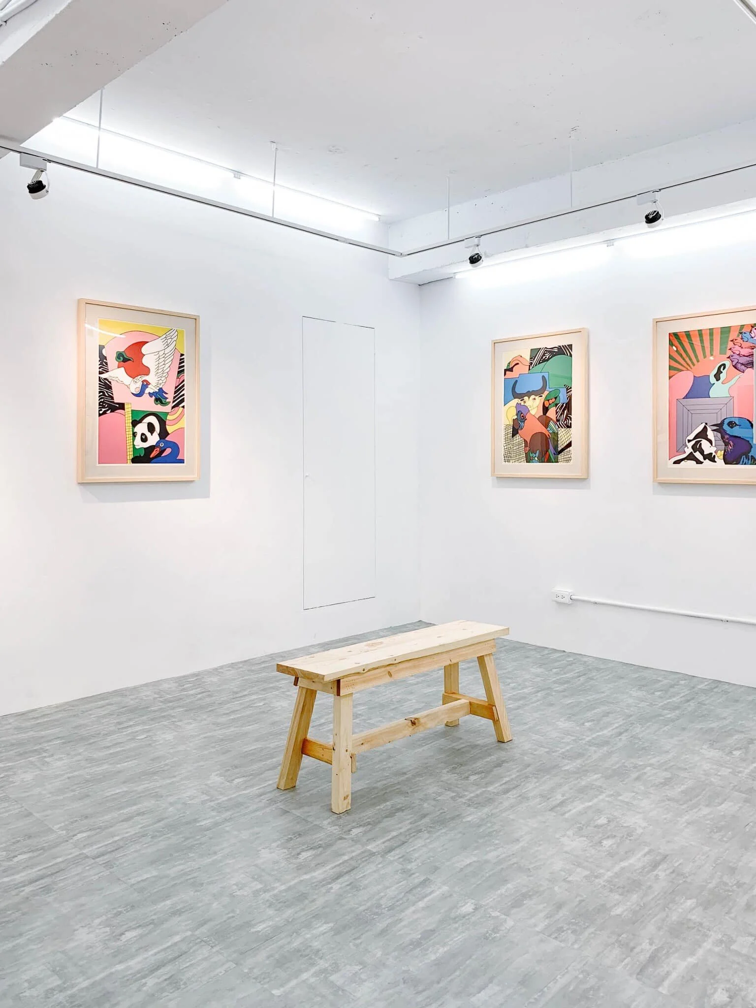 An art gallery with three framed colorful artworks on a white wall, a simple wooden bench in the center, and a gray floor.