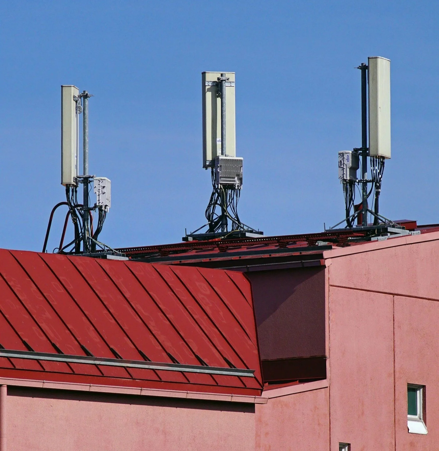 Three antennas or signal transmitters mounted on the roof of a pink building with a red roof and a clear blue sky in the background.