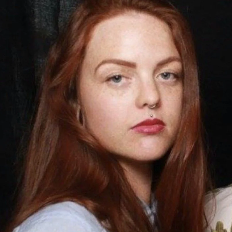 Close-up of a woman with long red hair, fair skin, blue eyes, and a nose and labret piercing, wearing a white top, against a dark background.