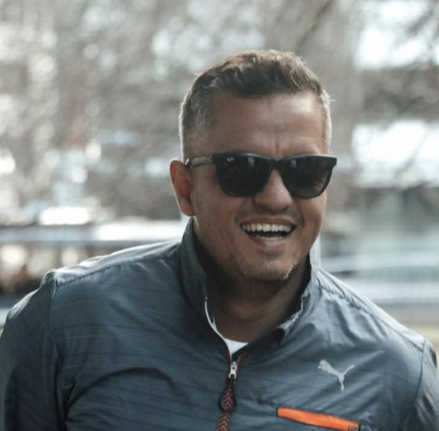 A man with short hair, wearing sunglasses and a dark blue jacket, smiling outdoors.