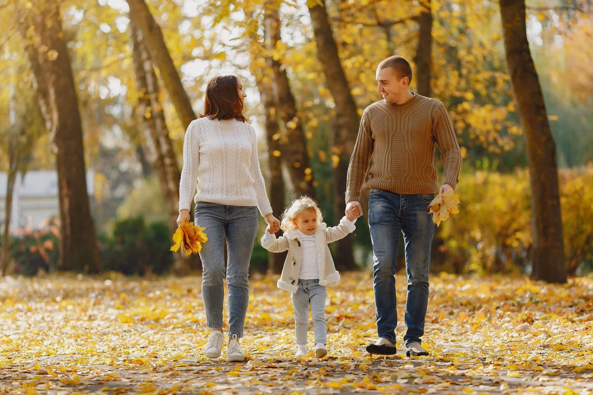 Two parents walk with their child through the woods while collecting fall leaves.