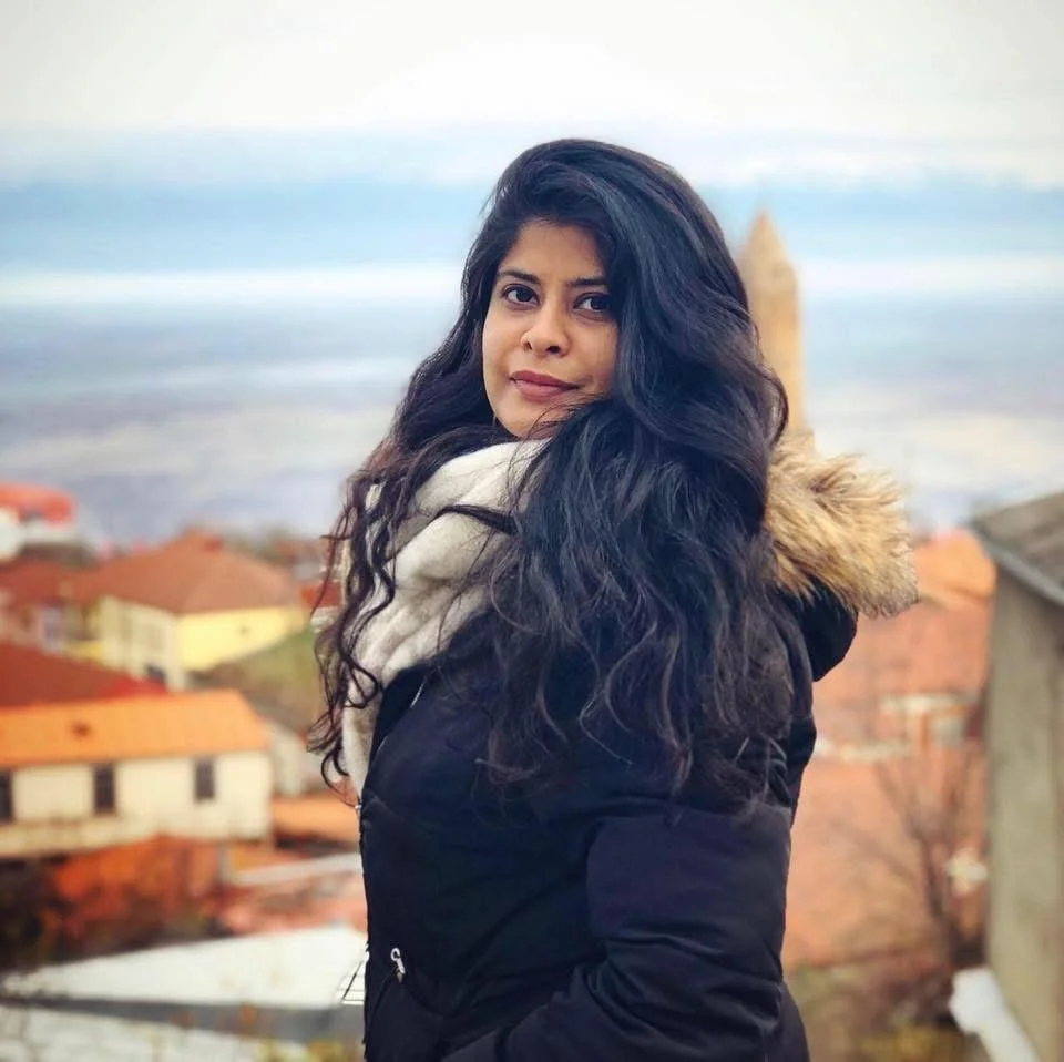 A woman with long, wavy black hair standing outdoors on a hill with a view of a coastal village and the ocean in the background.
