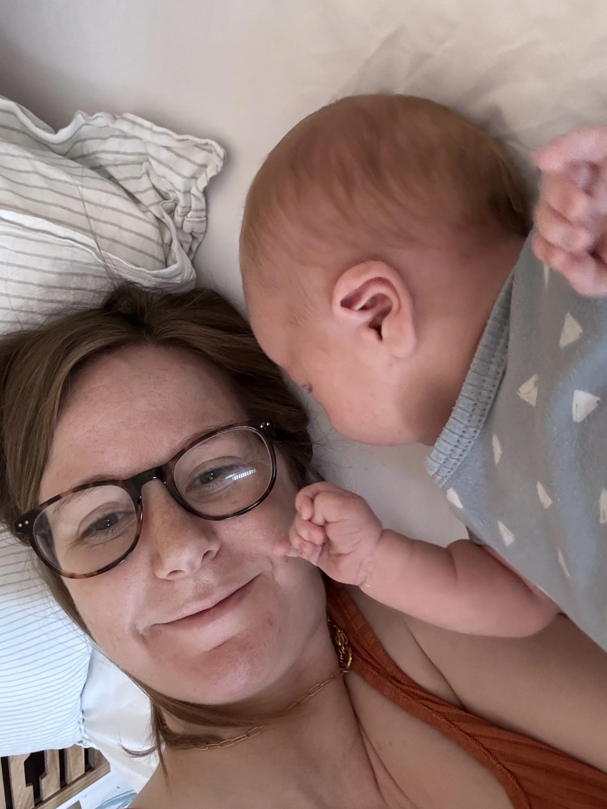 A woman with glasses and brown hair lying on a bed next to a baby with light brown hair, who is touching her face.