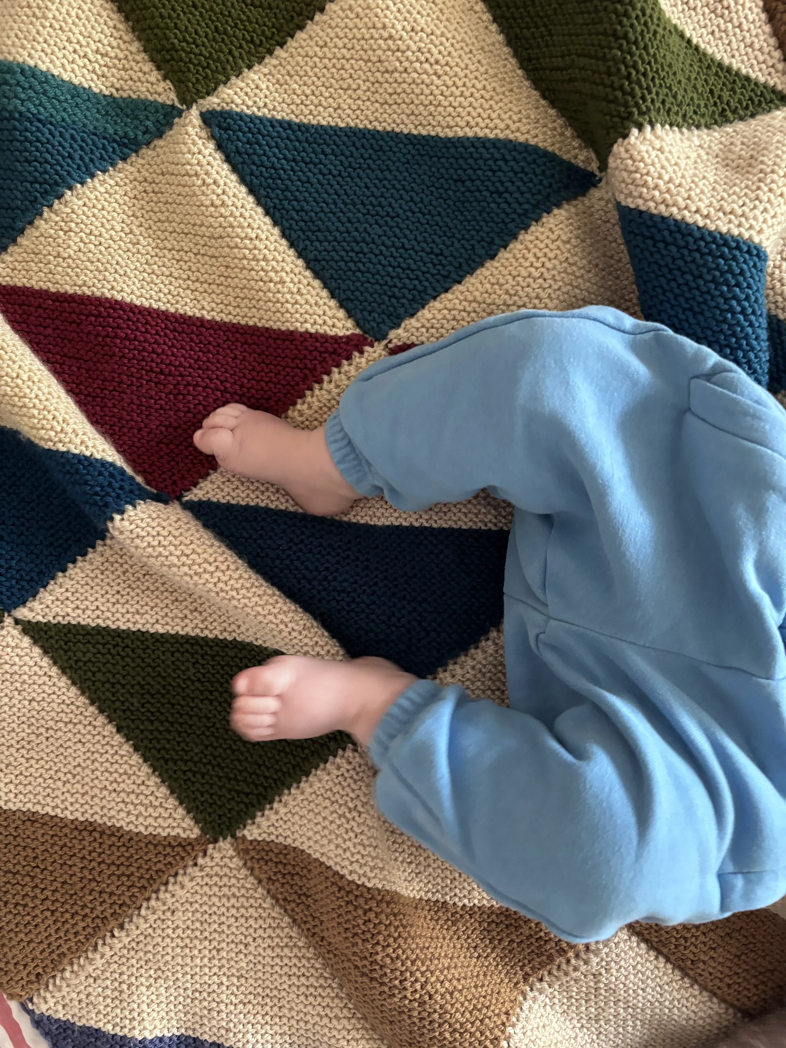 Close-up of a baby's feet with toes visible, wearing light blue joggers, lying on a geometric knitted blanket