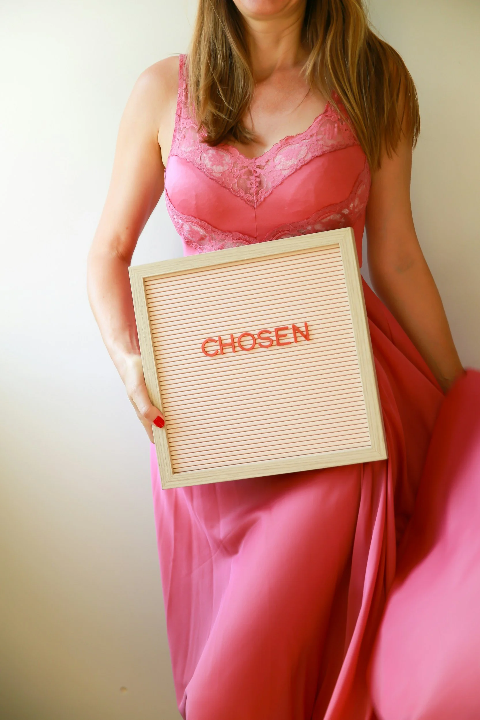 A woman in a pink lace dress holding a letter board with the word 'CHOSEN' written on it, standing against a plain beige background.