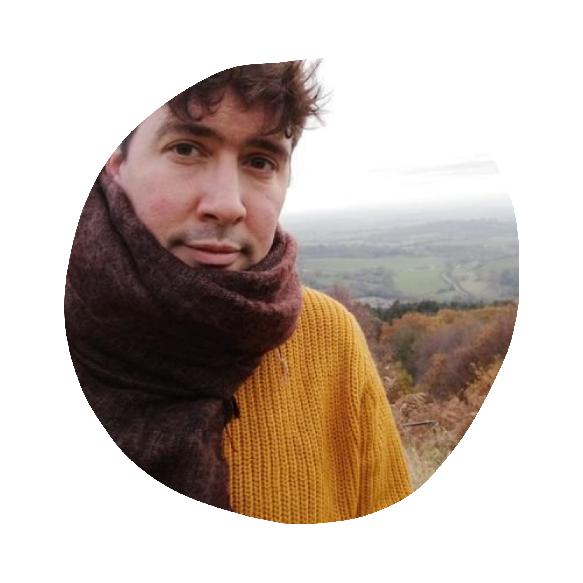 A person with curly hair smiling outdoors, wearing a mustard yellow sweater and a maroon scarf, with a landscape of fields and distant mountains in the background.