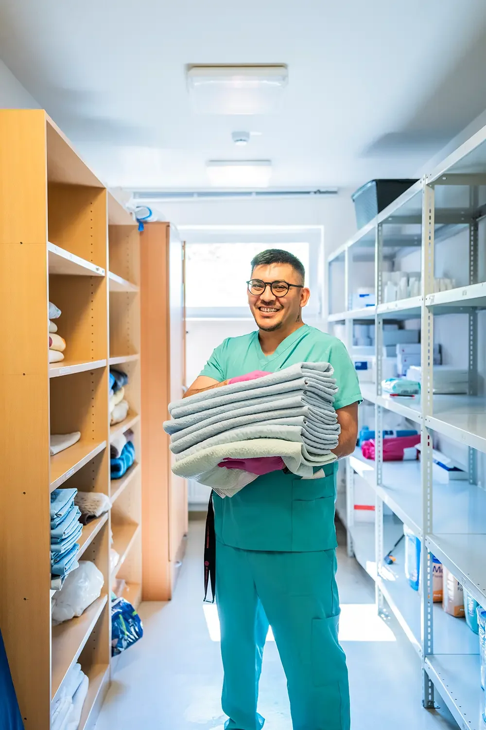 Ein Mann in grüner medizinischer Kleidung hält einen Stapel saubere Handtücher in einem Lagerraum. Der Raum ist mit Holz- und Metallregalen gefüllt, die verschiedene Textilien und medizinische Utensilien enthalten.