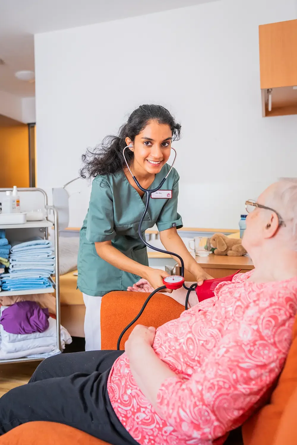 Eine junge Krankenschwester mit einem Stethoskop, die älterer Patientin im Pflegeheim die Blutdruck misst.