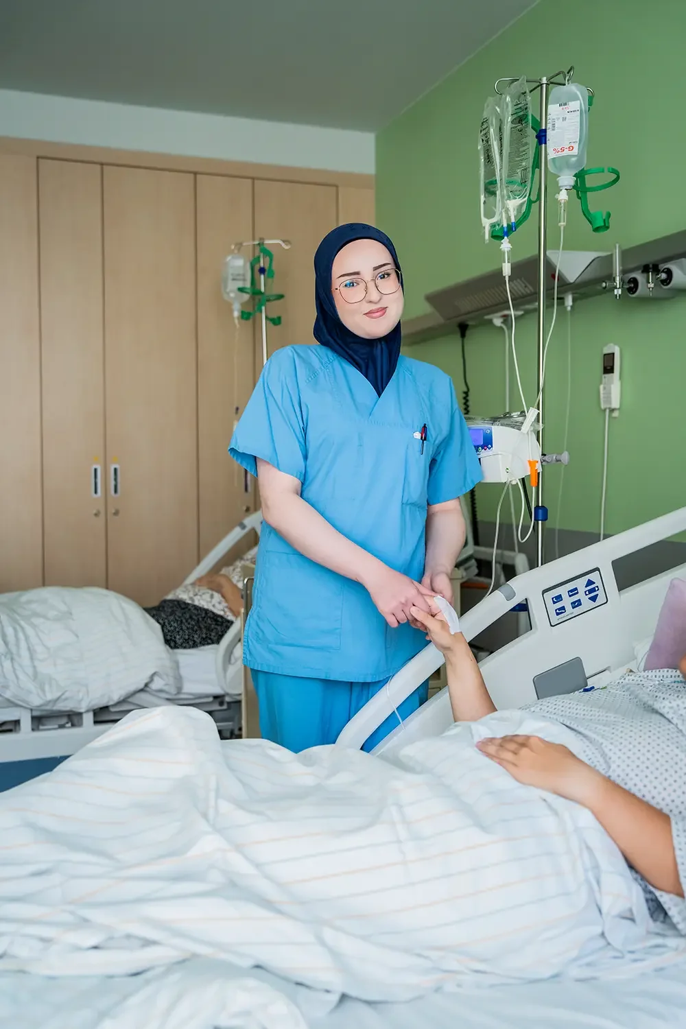 Empathische Krankenschwester hält Hand eines Patienten im Krankenhausbett, im Hintergrund medizinische Geräte und ein Patient, der auf einer Liege liegt.