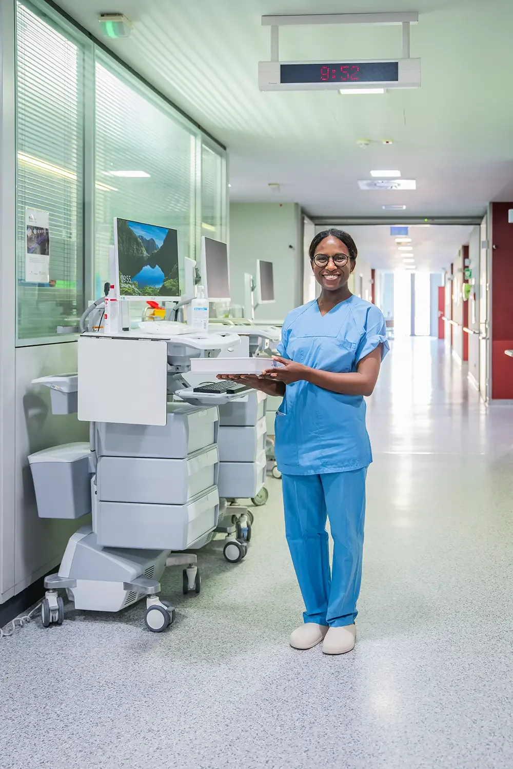 Ein lächelnder Arzt oder Krankenschwester in blauer medizinischer Kleidung steht in einem Klinikflur neben einem medizinischen Gerät mit einem Computerbildschirm, in einem gut beleuchteten Krankenhaus oder Klinik.