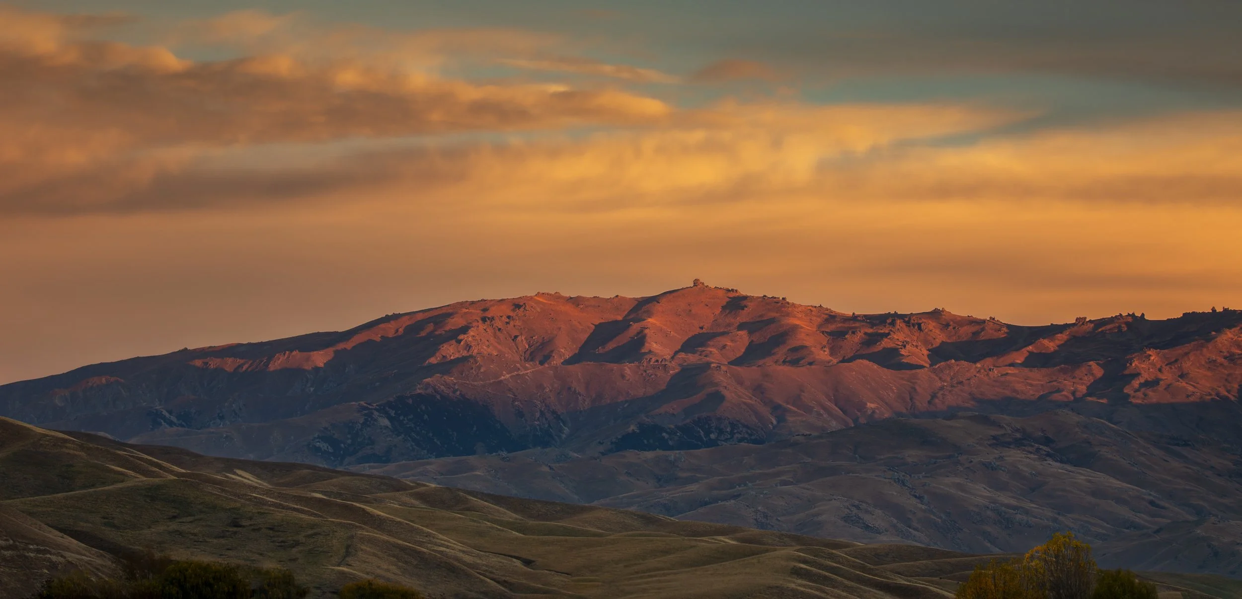 Sunset over a mountain range with orange and pink hues in the sky, rolling hills in the foreground.