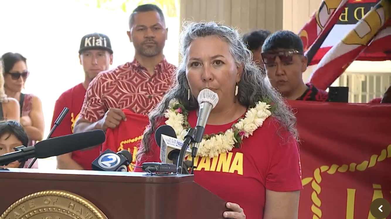 A woman at a podium speaking into a microphone, wearing a red shirt and a lei, with a group of people behind her, some holding a red and yellow banner.