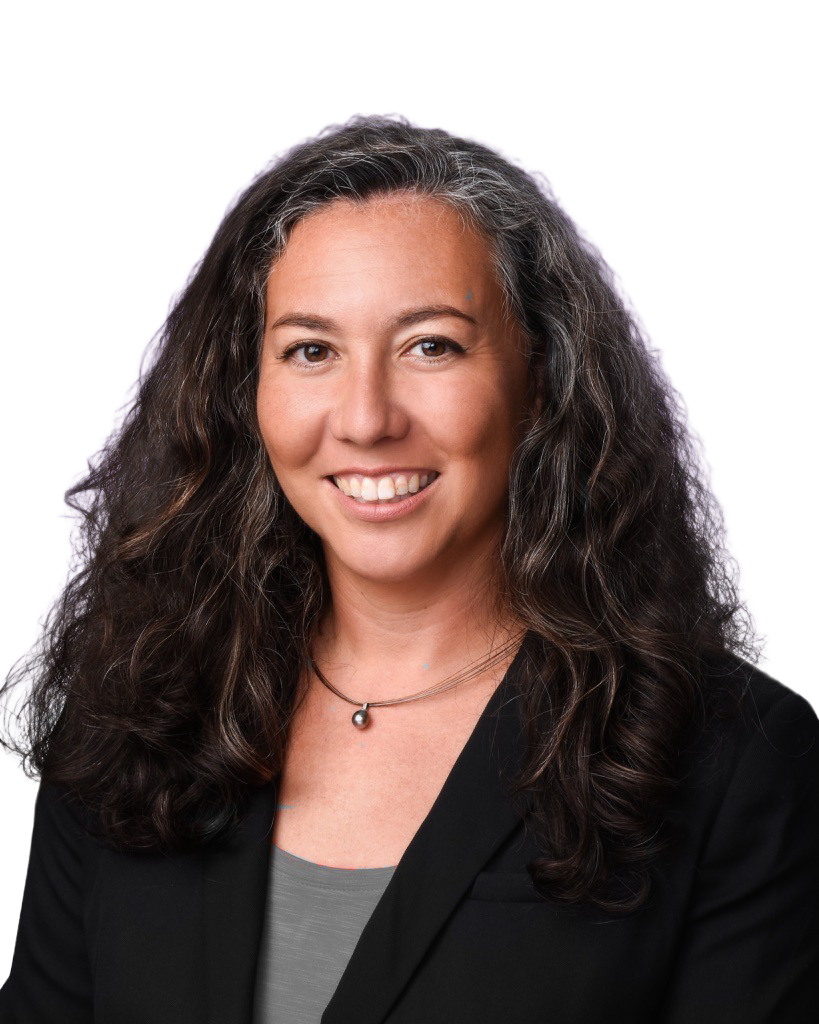 A woman with long, curly hair smiling, wearing a black blazer and a gray top, with a small necklace and a purple background.