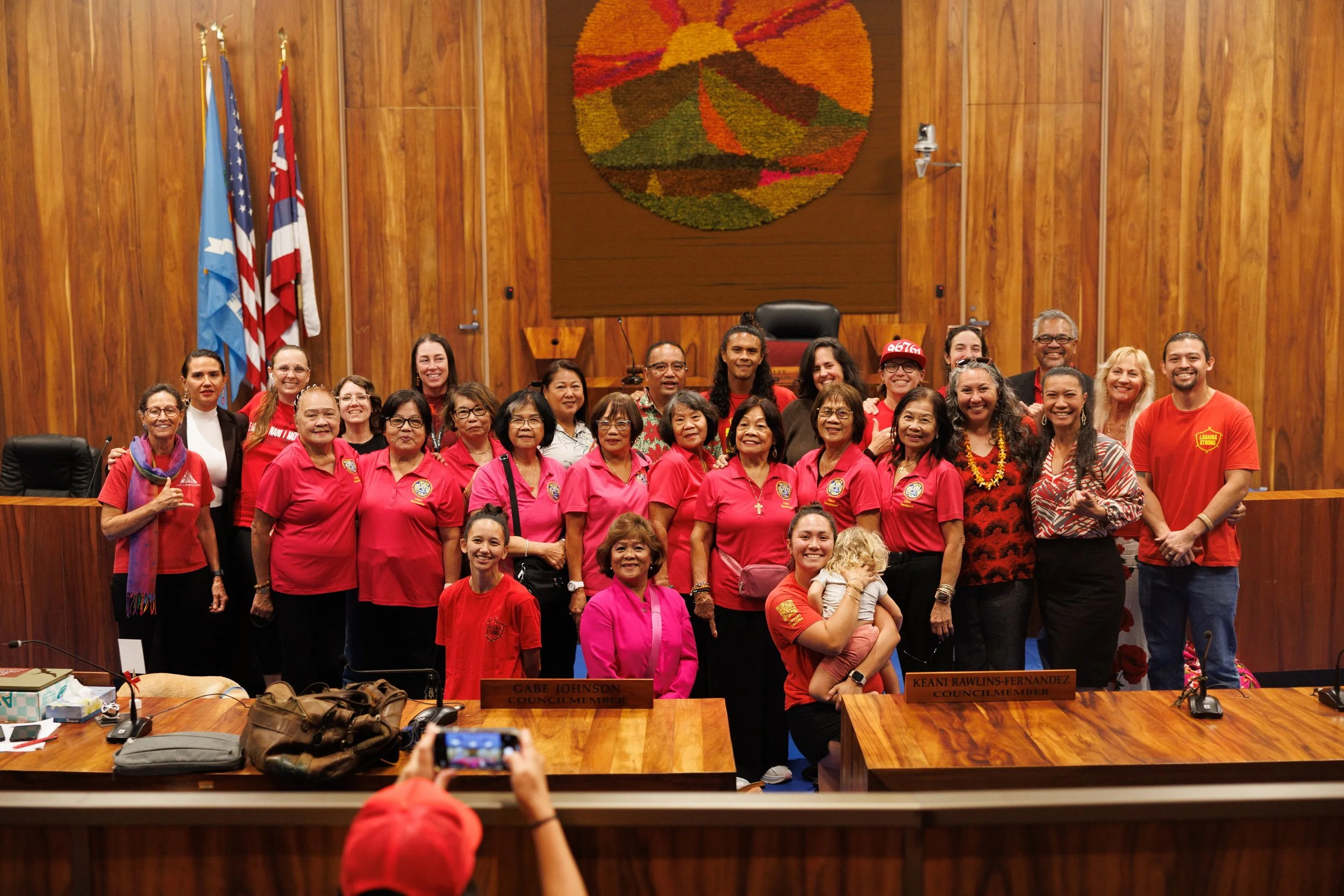 Group of diverse people, including women and men, smiling and gathered inside a wood-paneled room with flags and a large colorful wall hanging in the background. Some individuals wear bright pink shirts; others wear casual or formal attire. A person in the foreground holds up a smartphone, capturing the moment.