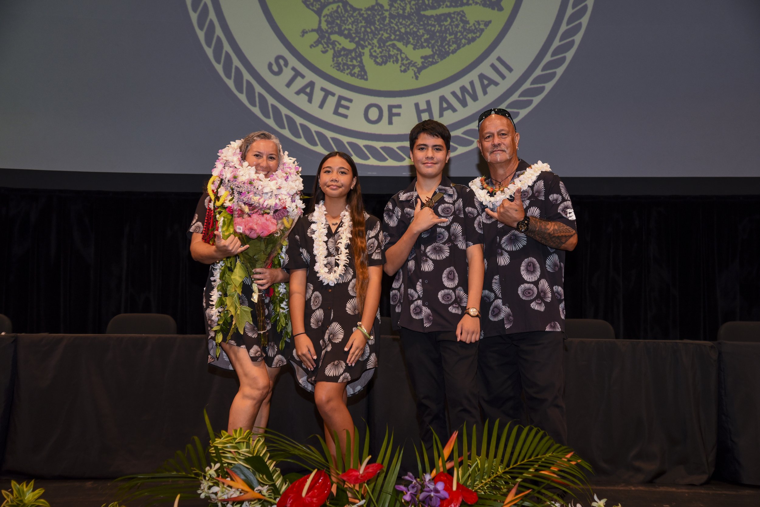 Group of four people on stage at an award ceremony, with a large emblem of the State of Hawaii projected behind them. The woman on the left is holding a bouquet of flowers and wearing a floral lei. The other three are wearing matching black Hawaiian shirts with white patterns. They are all smiling and making hand gestures, with flowers displayed at the front of the stage.