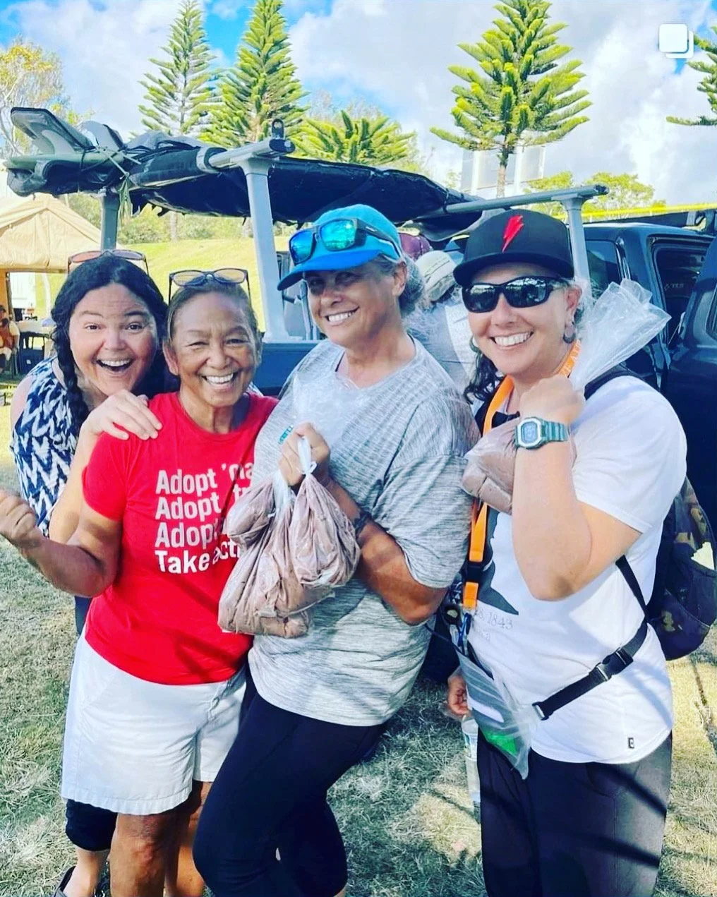Four women standing outdoors near a vehicle, smiling, with trees and a tent in the background, wearing casual clothes and accessories.