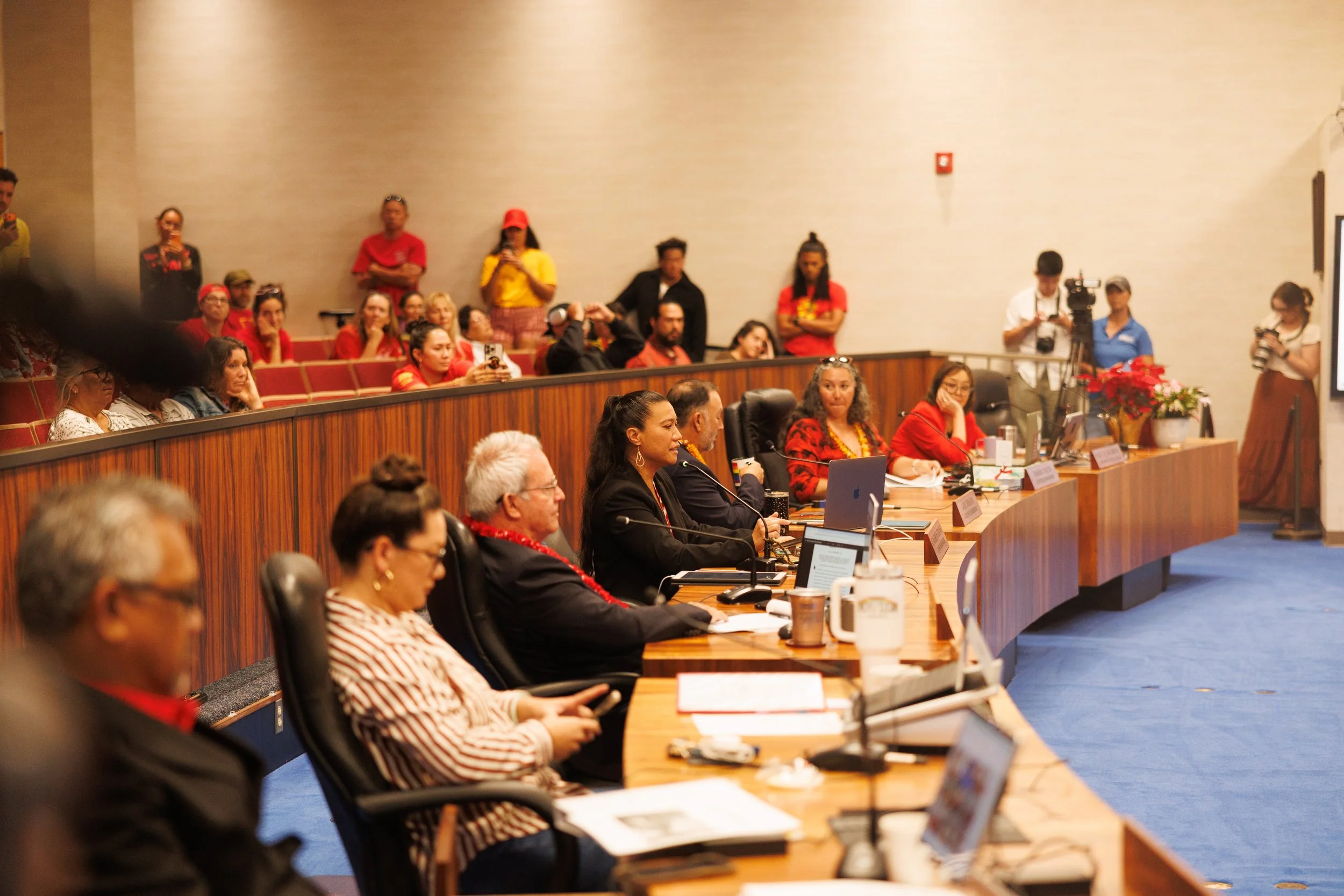 People seated at a long conference table with laptops, microphones, and documents, participating in a formal meeting or conference. Some people are taking notes or listening, while others are using phones or cameras. In the background, there are additional attendees standing or sitting against a wall, some taking pictures.