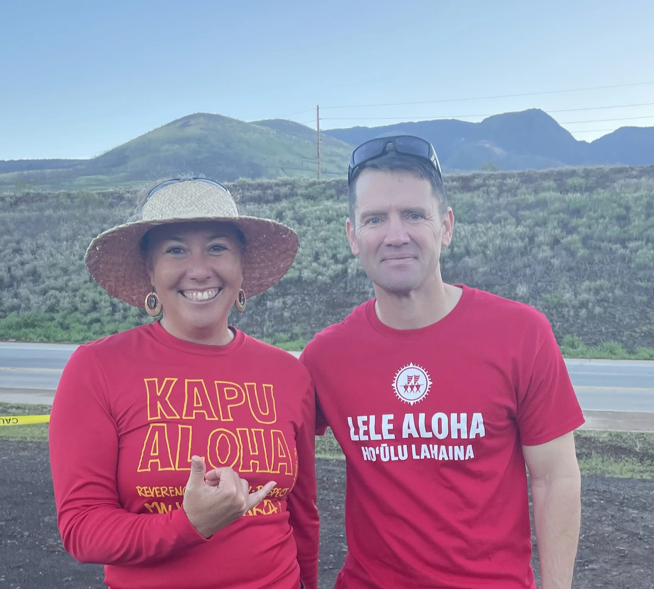 Two people wearing red shirts with yellow and white text on them, standing outdoors with hills and mountains in the background. One person is wearing a large sunhat and earrings, and the other has sunglasses on top of their head.