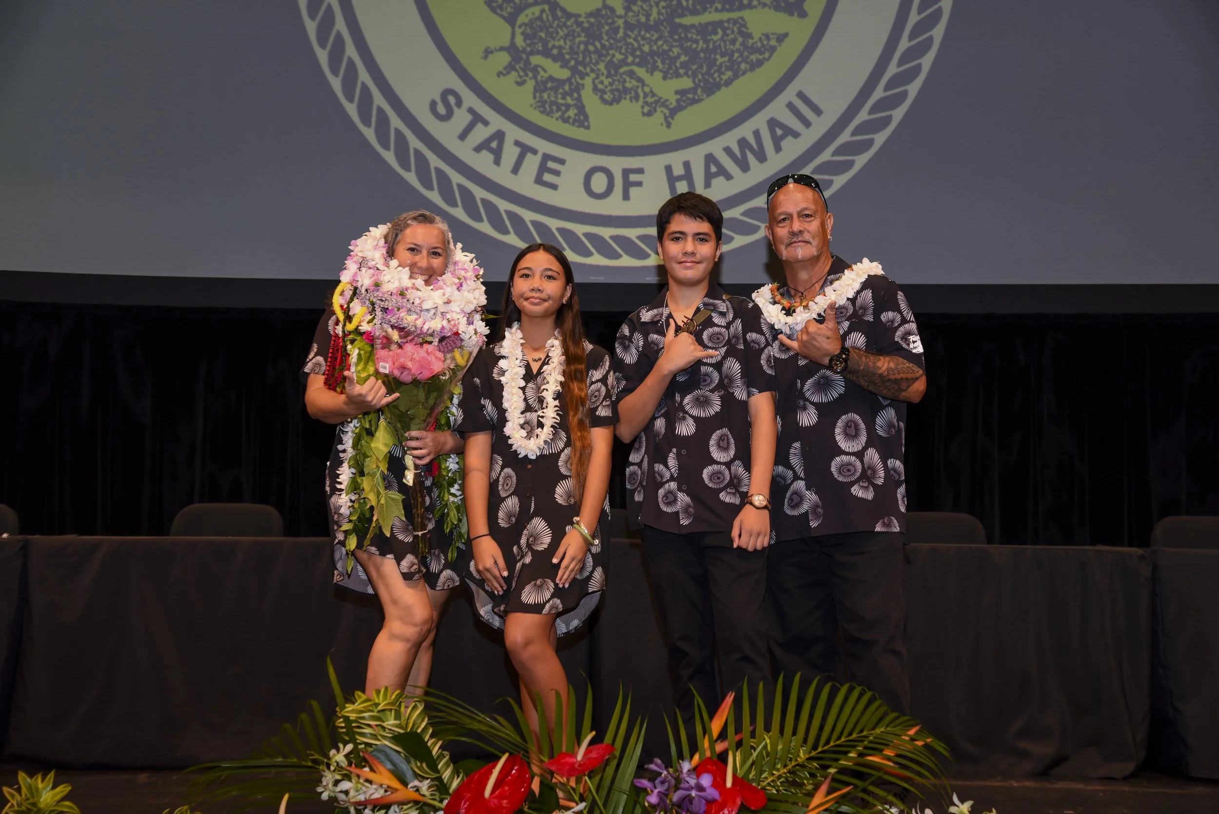 A group of four people standing on a stage with a large emblem of the State of Hawaii projected behind them. The group consists of two women and two young men, all wearing matching black Hawaiian shirts with white flower patterns and leis. The woman on the left holds a bouquet of flowers, while the young people are making hand gestures with their right hands. There is a floral arrangement in front of the stage.