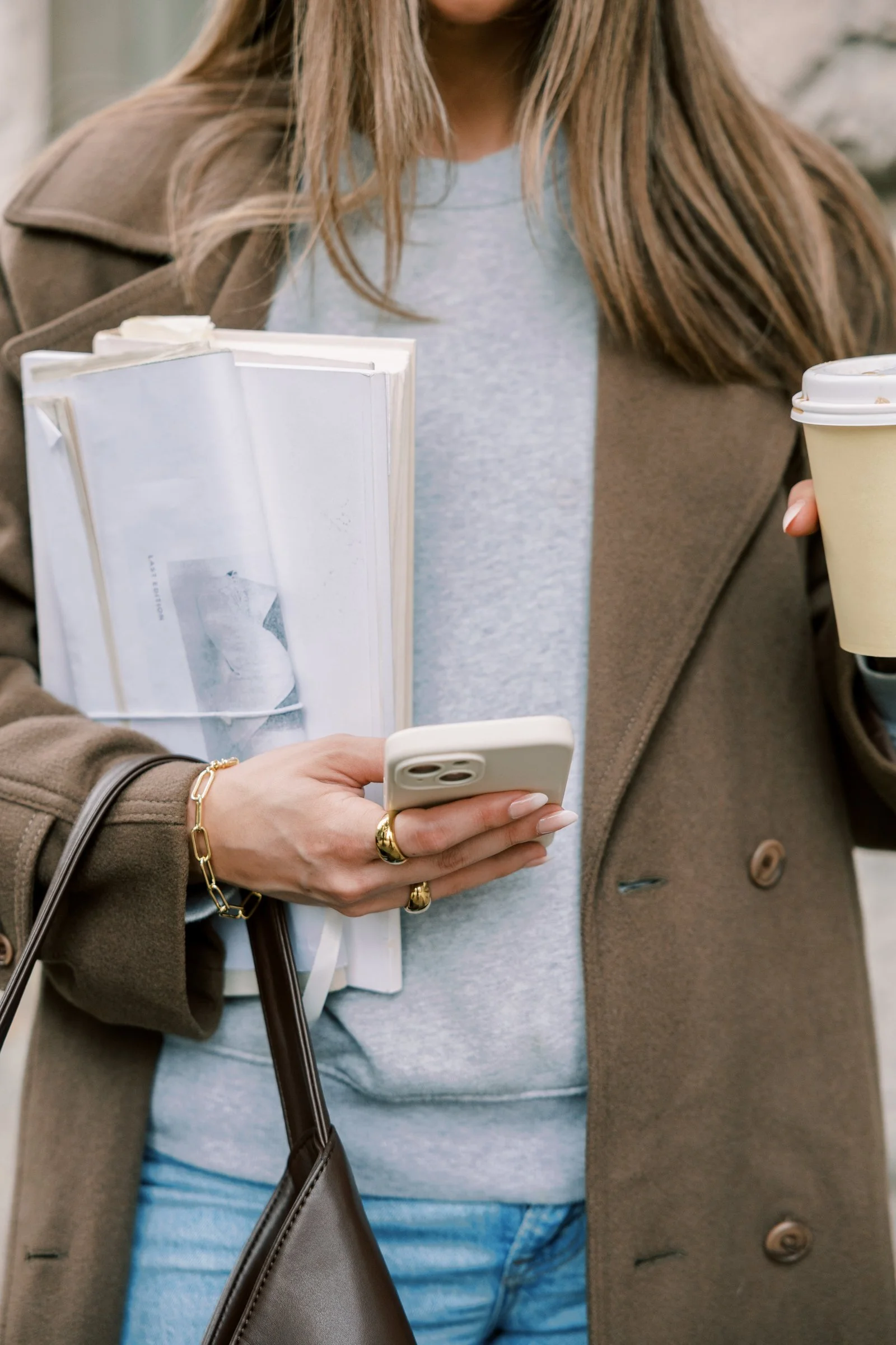 A woman in a beige overcoat, holding a disposable coffee cup, wearing a gray sweatshirt, gold jewelry, and carrying a brown purse, is looking at her phone.
