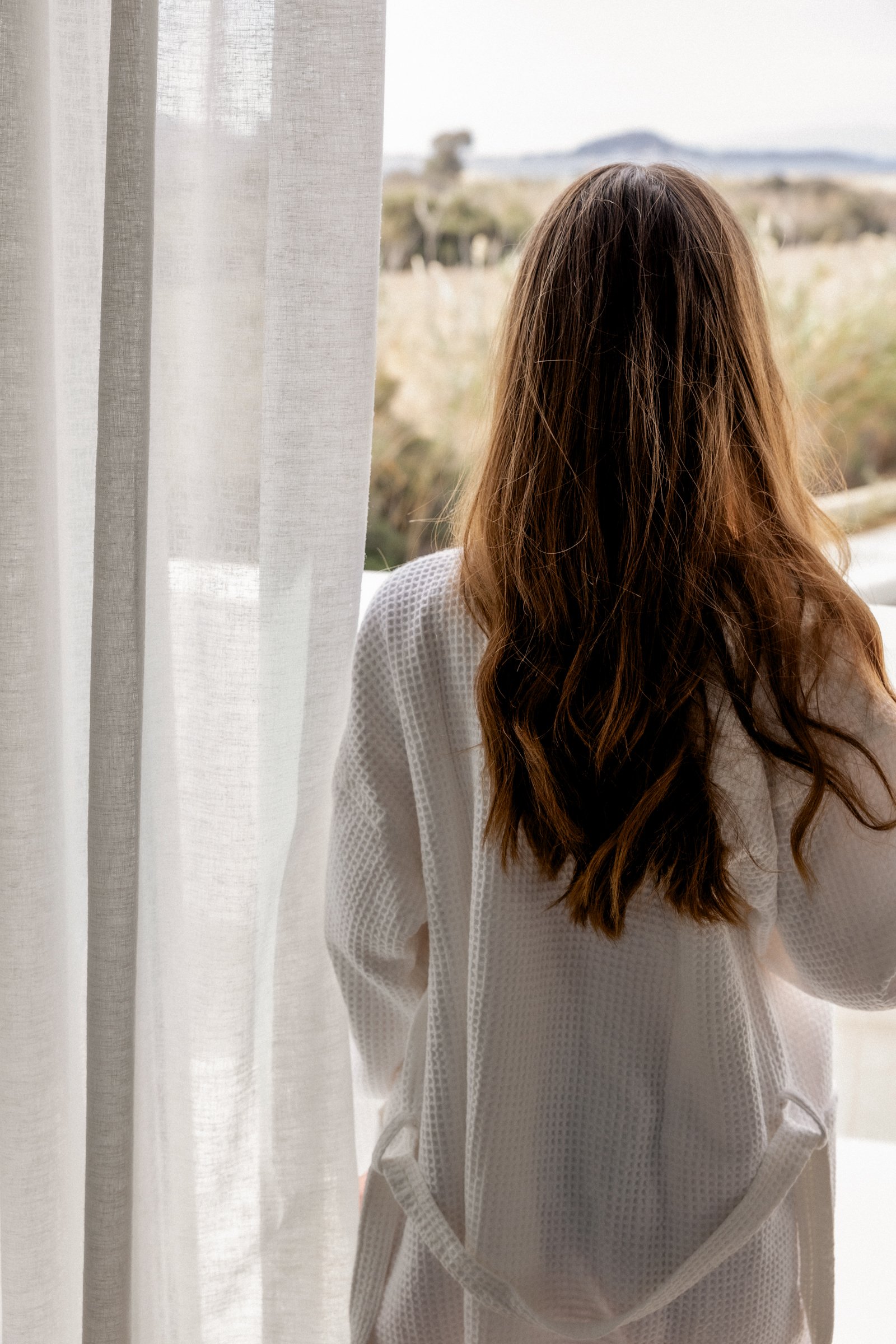A woman with long, wavy brown hair wearing a white bathrobe looking out a window with sheer white curtains, overlooking a landscape with trees and hills in the distance.