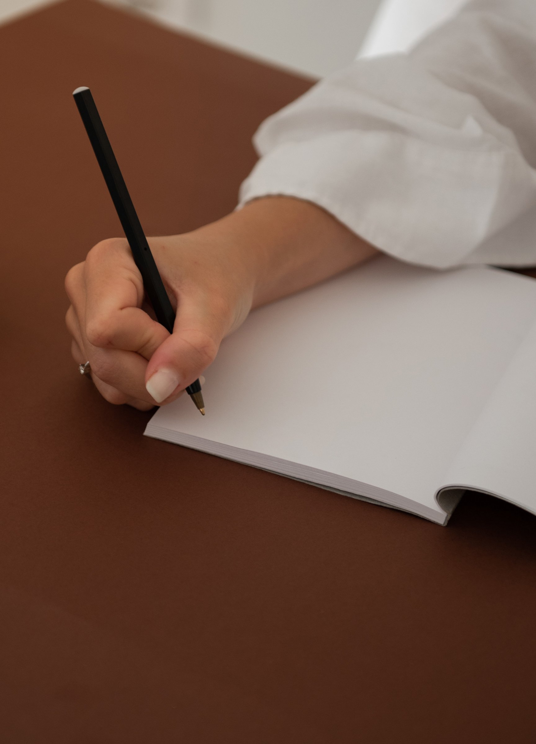 Close-up of a person in a white shirt writing on a notepad with a black pen on a brown desk.