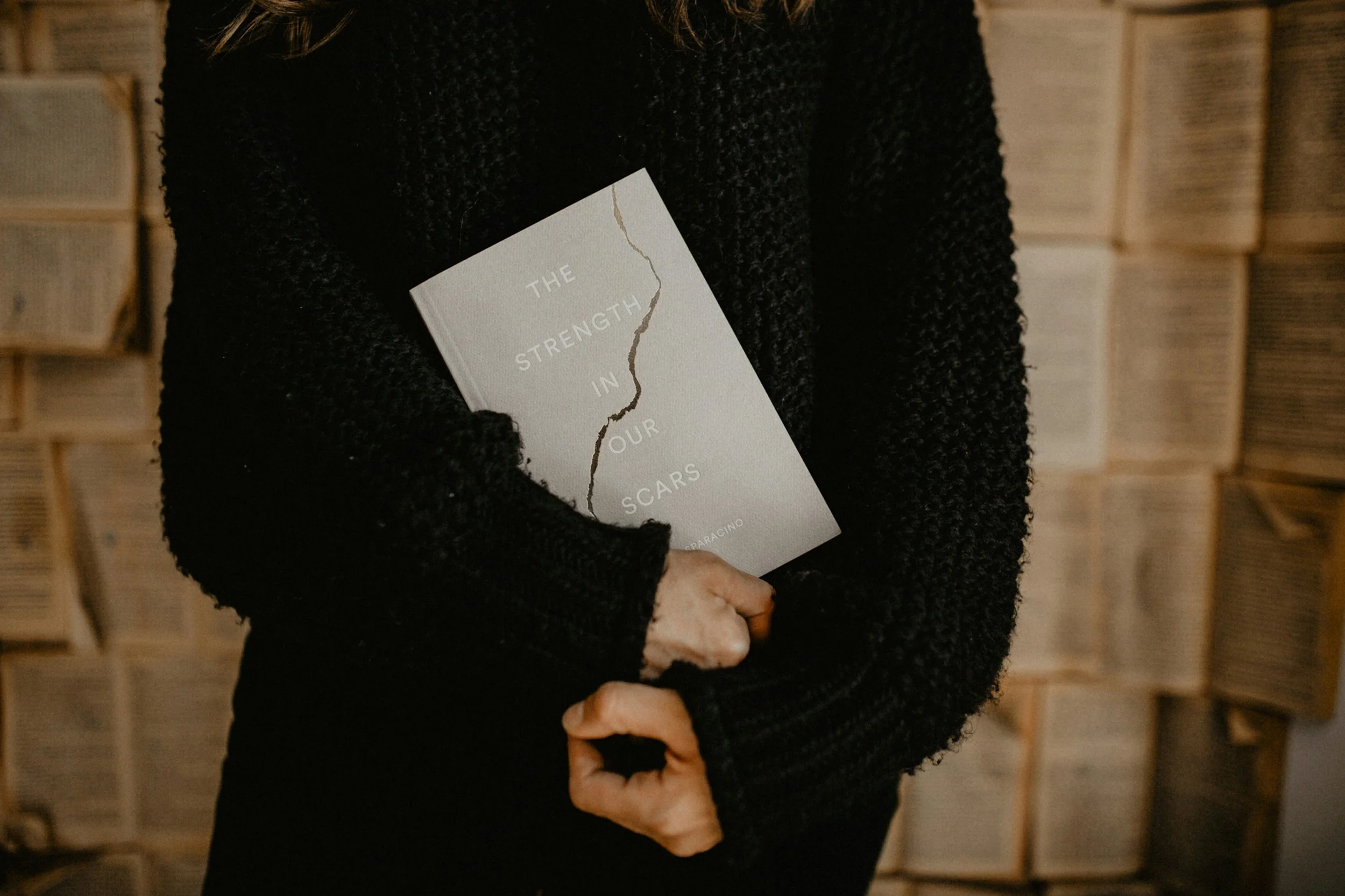 Person holding a book titled 'The Strength in Our Scars' in front of a wall of open books.
