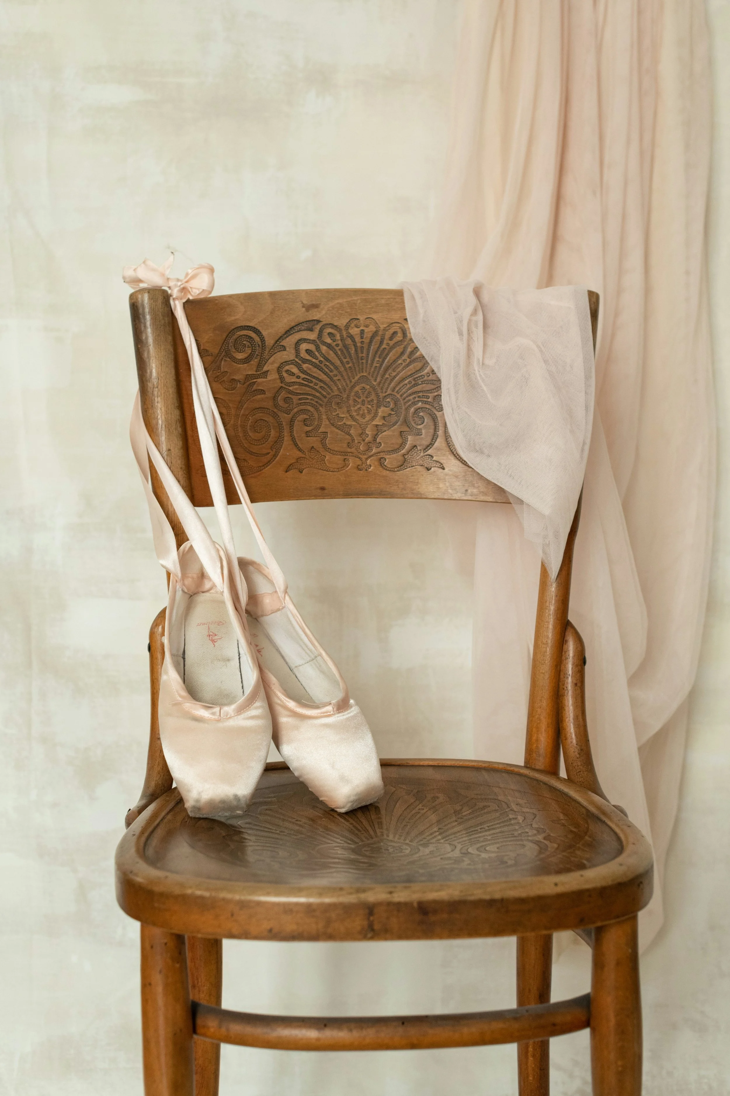 A wooden chair with ballet slippers hanging from the top left corner of the backrest and a sheer, light-colored fabric draped over the right side of the backrest.