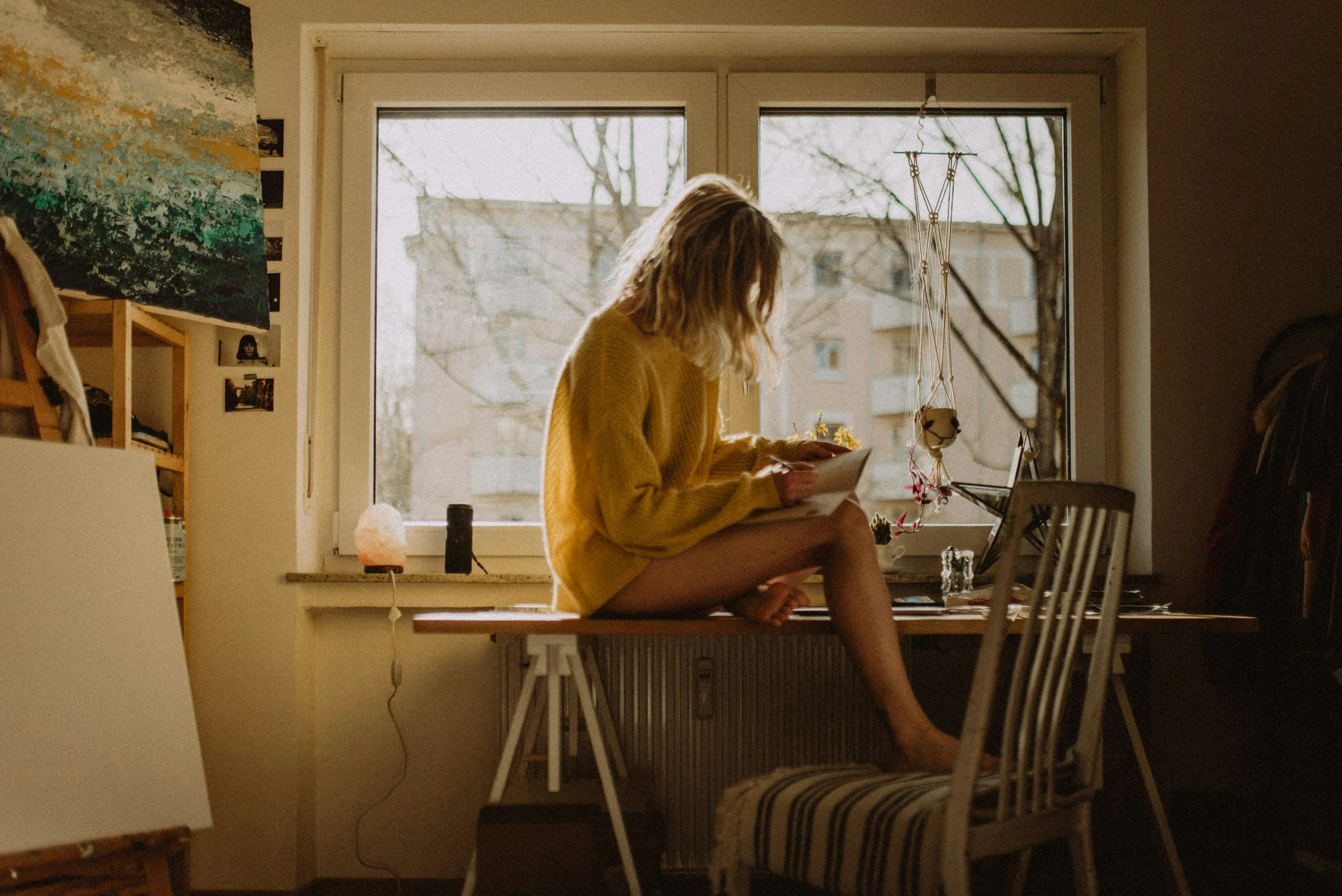 A woman with blonde hair, wearing a yellow oversized sweater, sitting cross-legged on a wooden table near a window, reading a book or notebook. The room has a cozy, artistic vibe with paintings on the wall, shelves with books and photos, a salt lamp, and some hanging decorations. Outside the window, there are leafless trees and residential buildings.