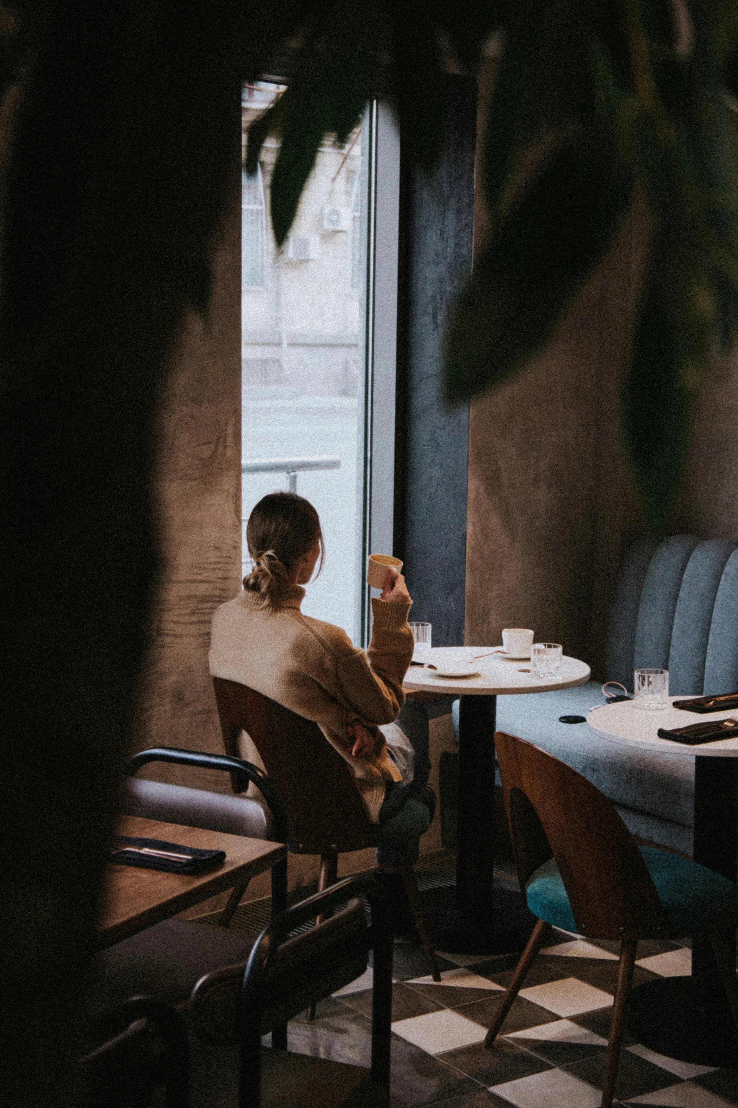 A woman sitting at a table in a dimly lit restaurant, holding a cup, with a window in the background revealing a blurred urban street scene outside.
