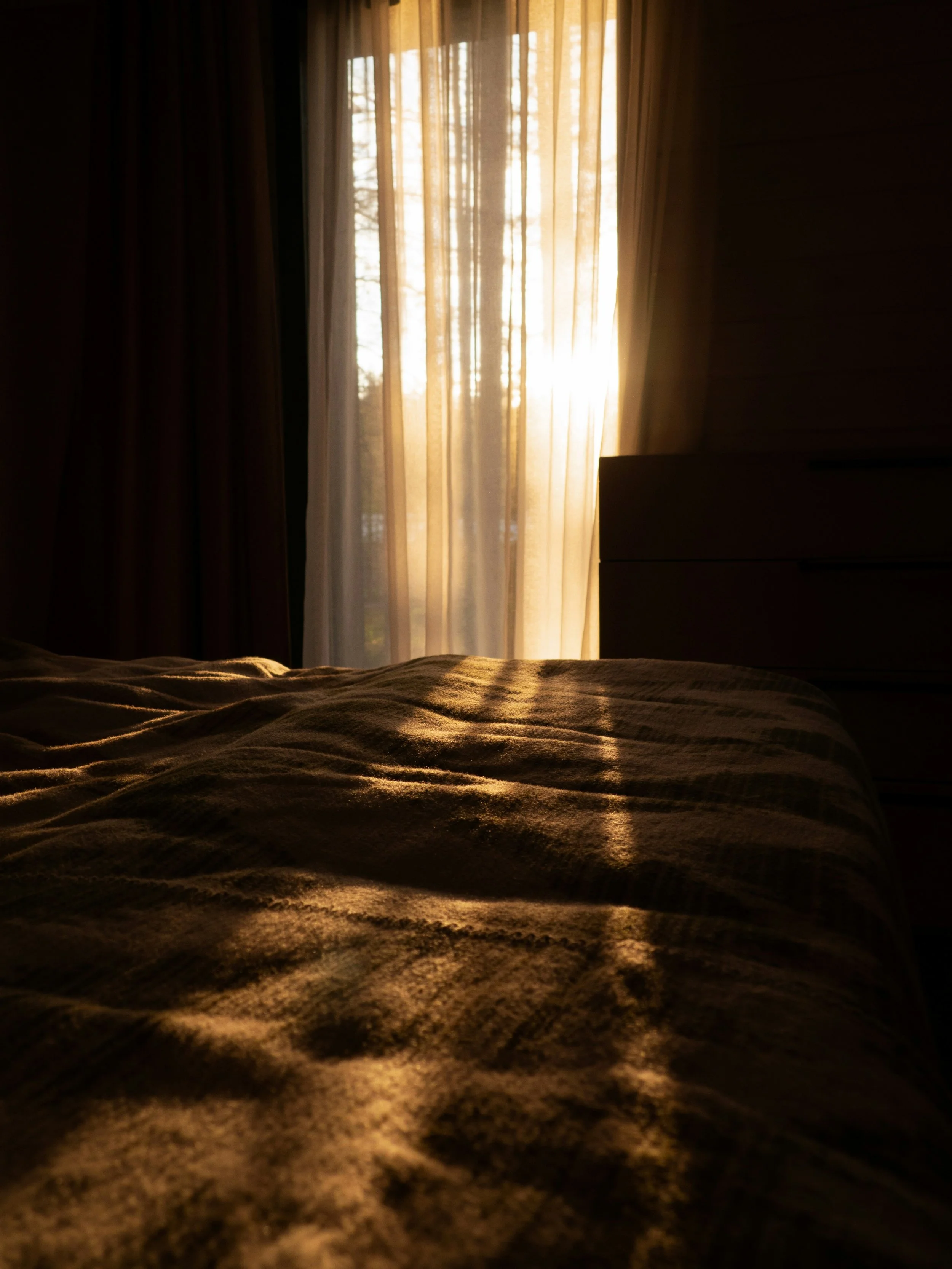 Sunlight coming through a window with beige curtains, illuminating a bedspread with a brown, textured pattern.