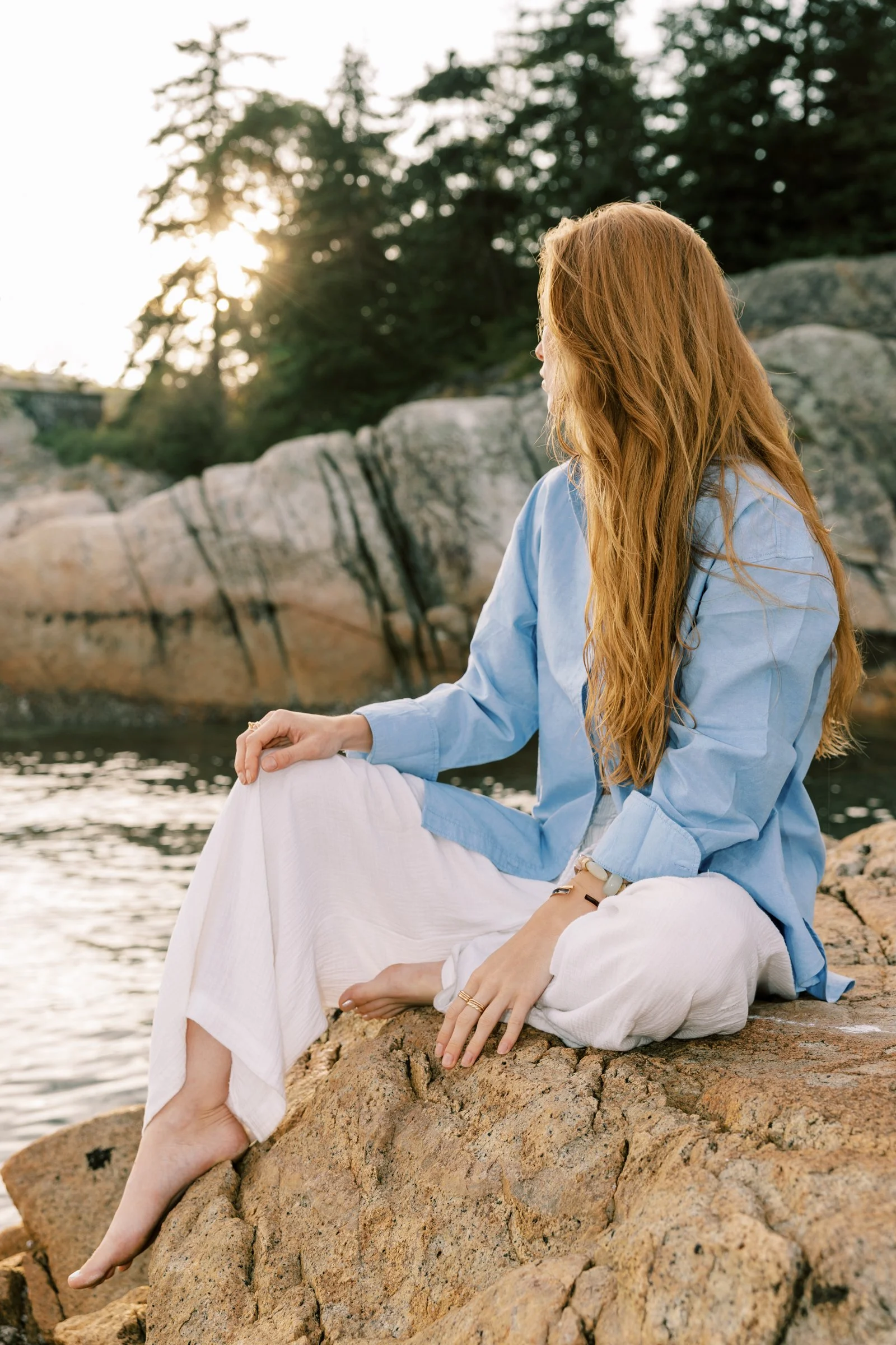 A woman with long red hair sitting on rocks near a body of water, wearing a blue shirt and white pants, with greenery and trees in the background, during sunset.